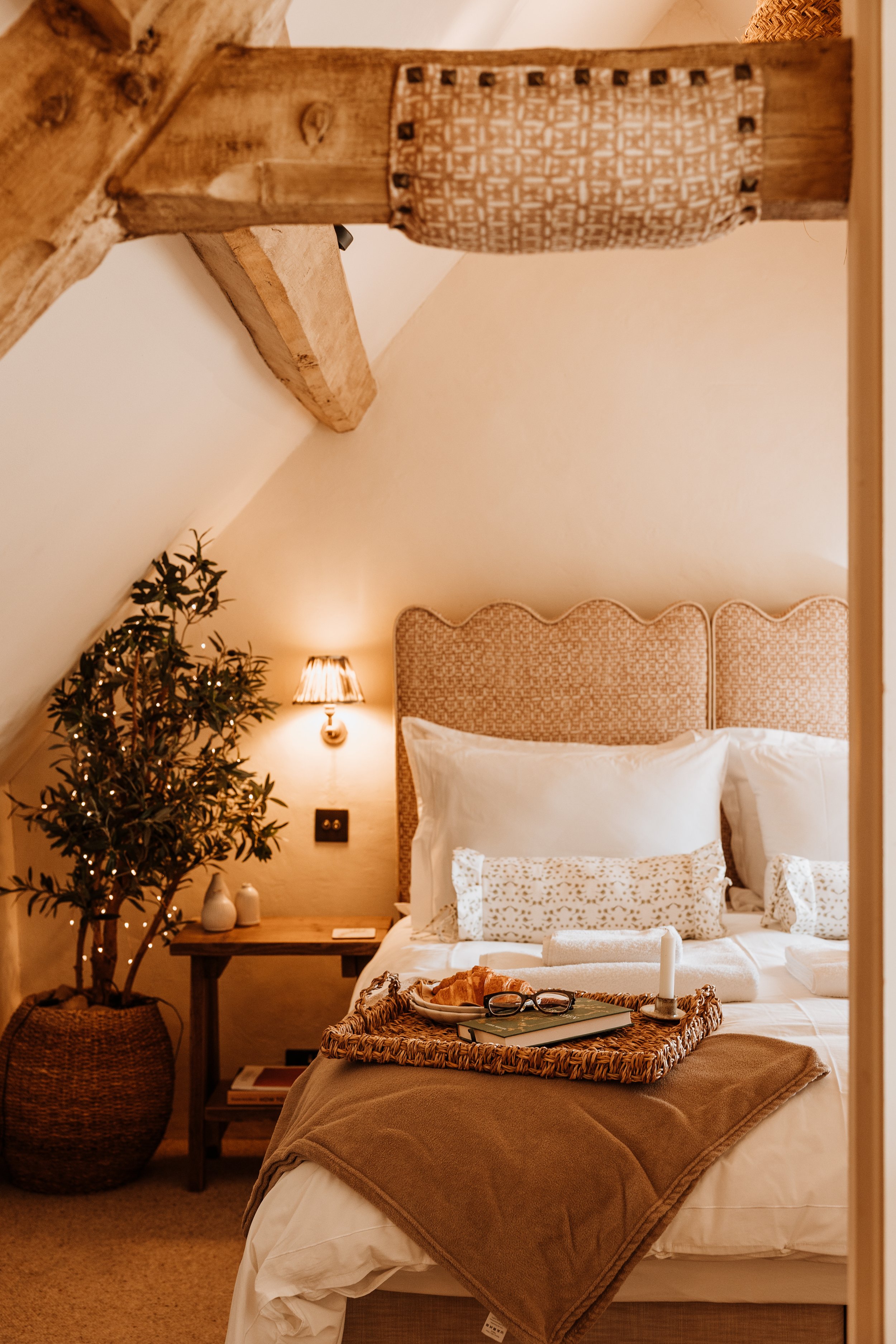 A cozy bedroom with an angled ceiling, featuring a bed with beige patterned headboard, white pillows, and a brown blanket. A tray with a croissant, glasses, a candle, and a book is on the bed.  Interiors by Lucy Rebecca Photography.