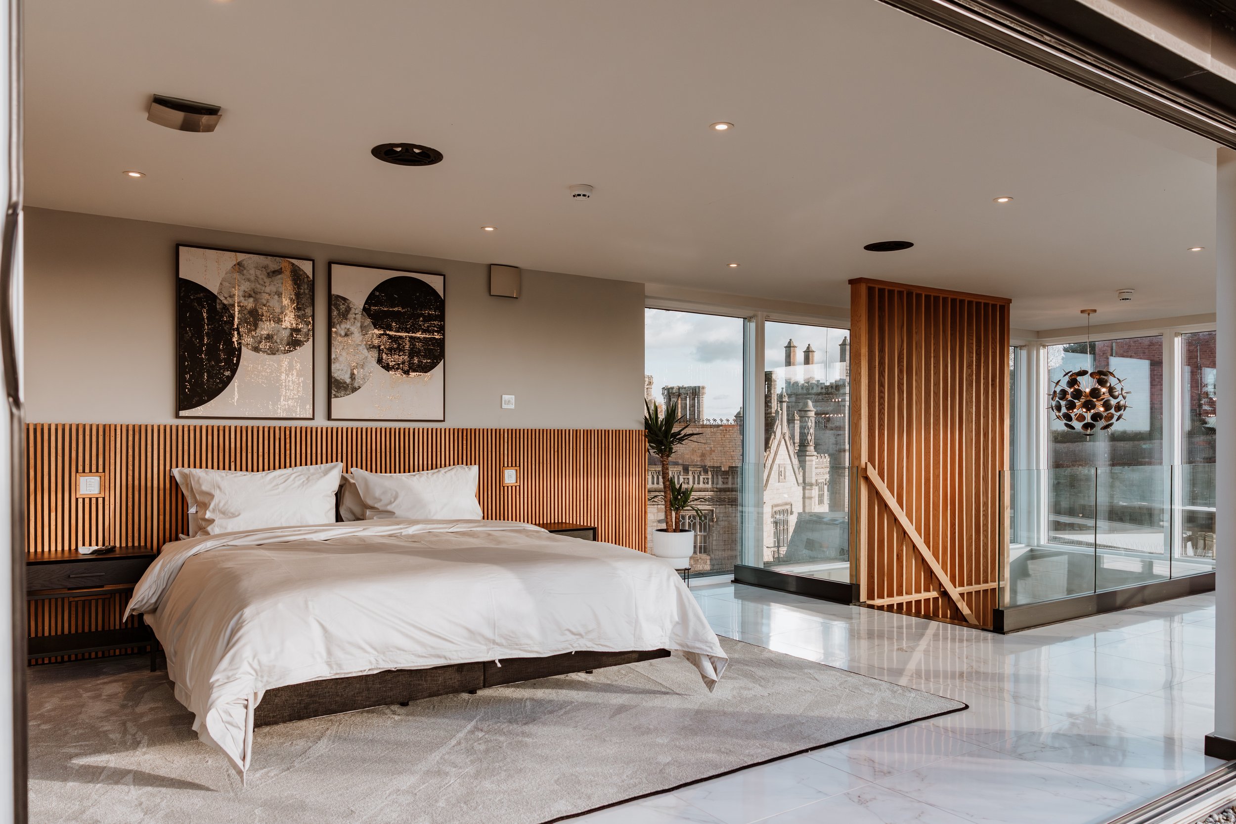 Modern bedroom with white bed, wooden headboard, abstract black and white artwork, potted plant, large windows with city view, and a glass railing balcony. Interiors by Lucy Rebecca Photography.