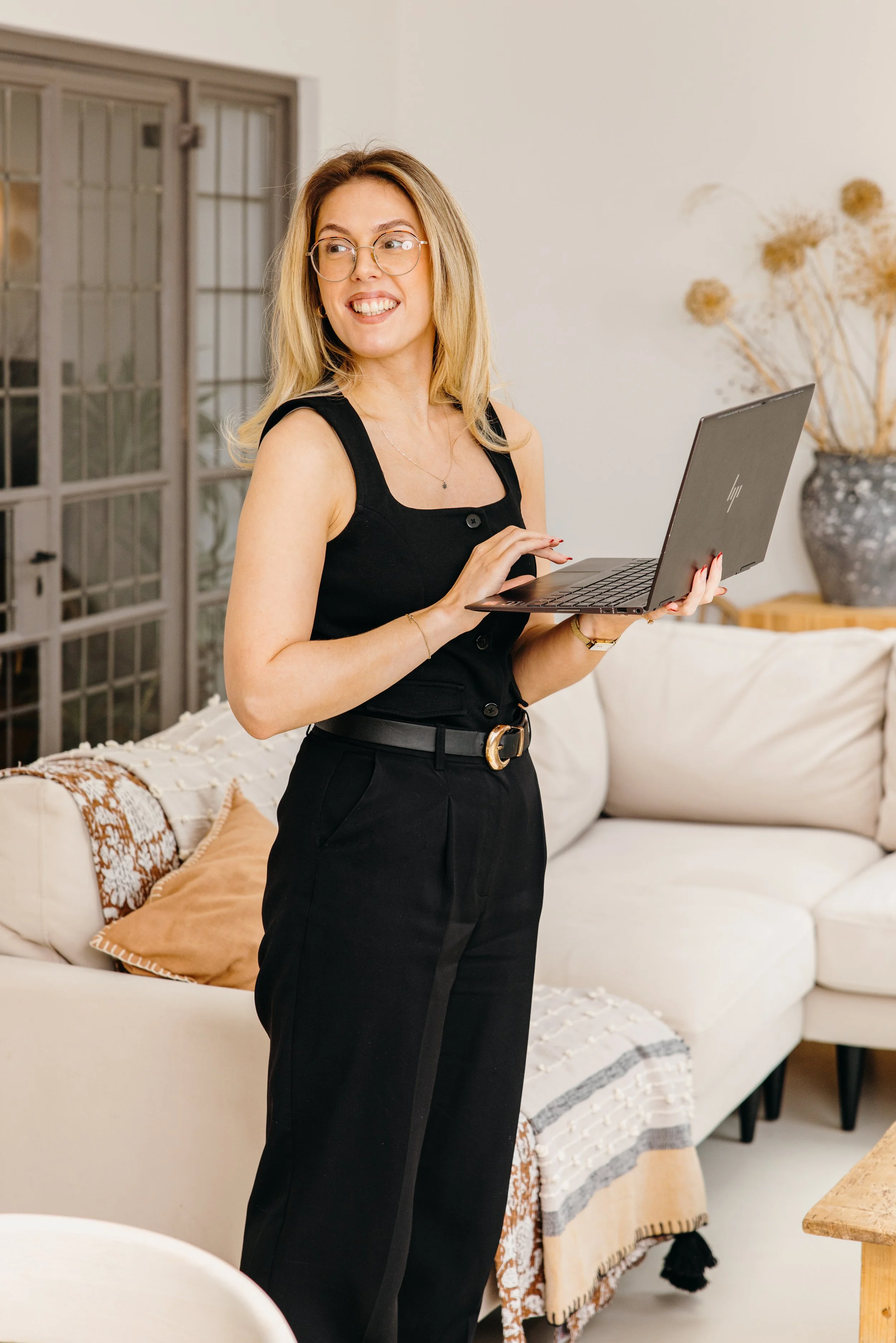 A woman with glasses and blonde hair, wearing a black sleeveless top and black pants, standing in a living room, smiling and holding a silver laptop. Personal Branding by Lucy Rebecca Photography.