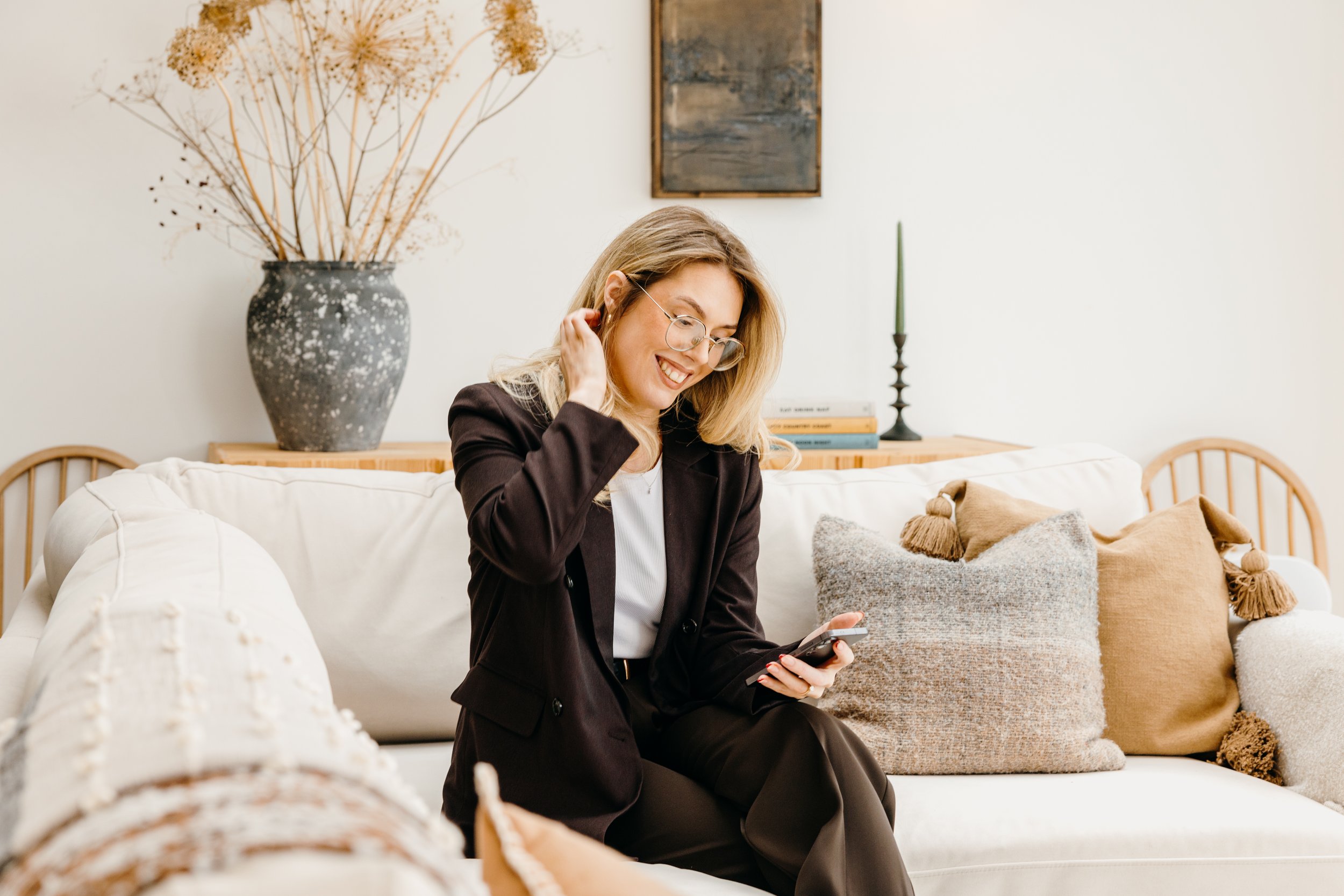 A woman sitting on a cream-colored sofa in a living room, smiling, while looking at her phone. She is wearing glasses, a black blazer, and black pants. Personal Branding by Lucy Rebecca Photography.