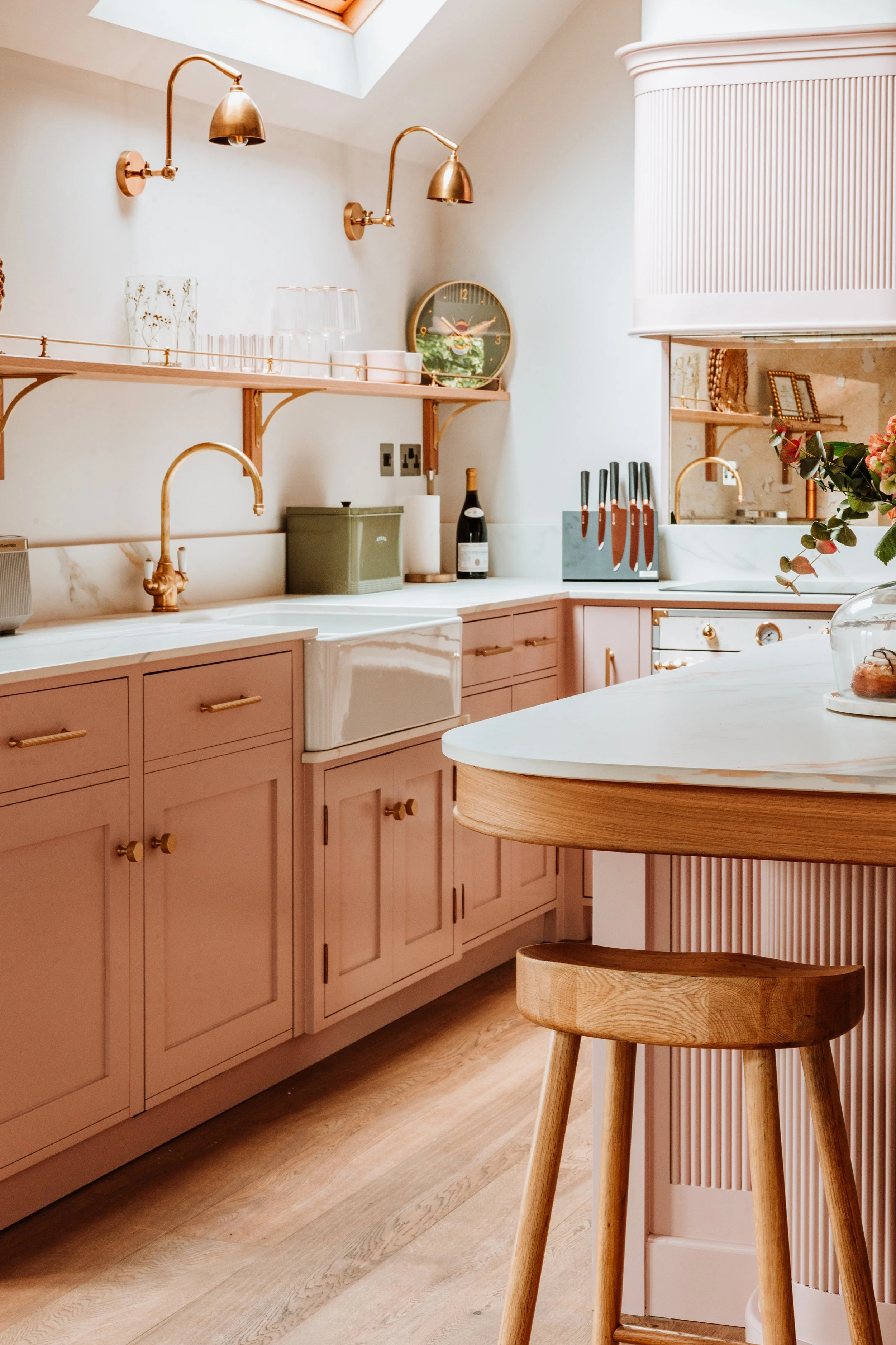 Modern kitchen with pink cabinets, white marble countertops, gold fixtures, and wooden accents. Skylight above, open shelving with glassware and decor, and a wooden stool. Interiors by Lucy Rebecca Photography.