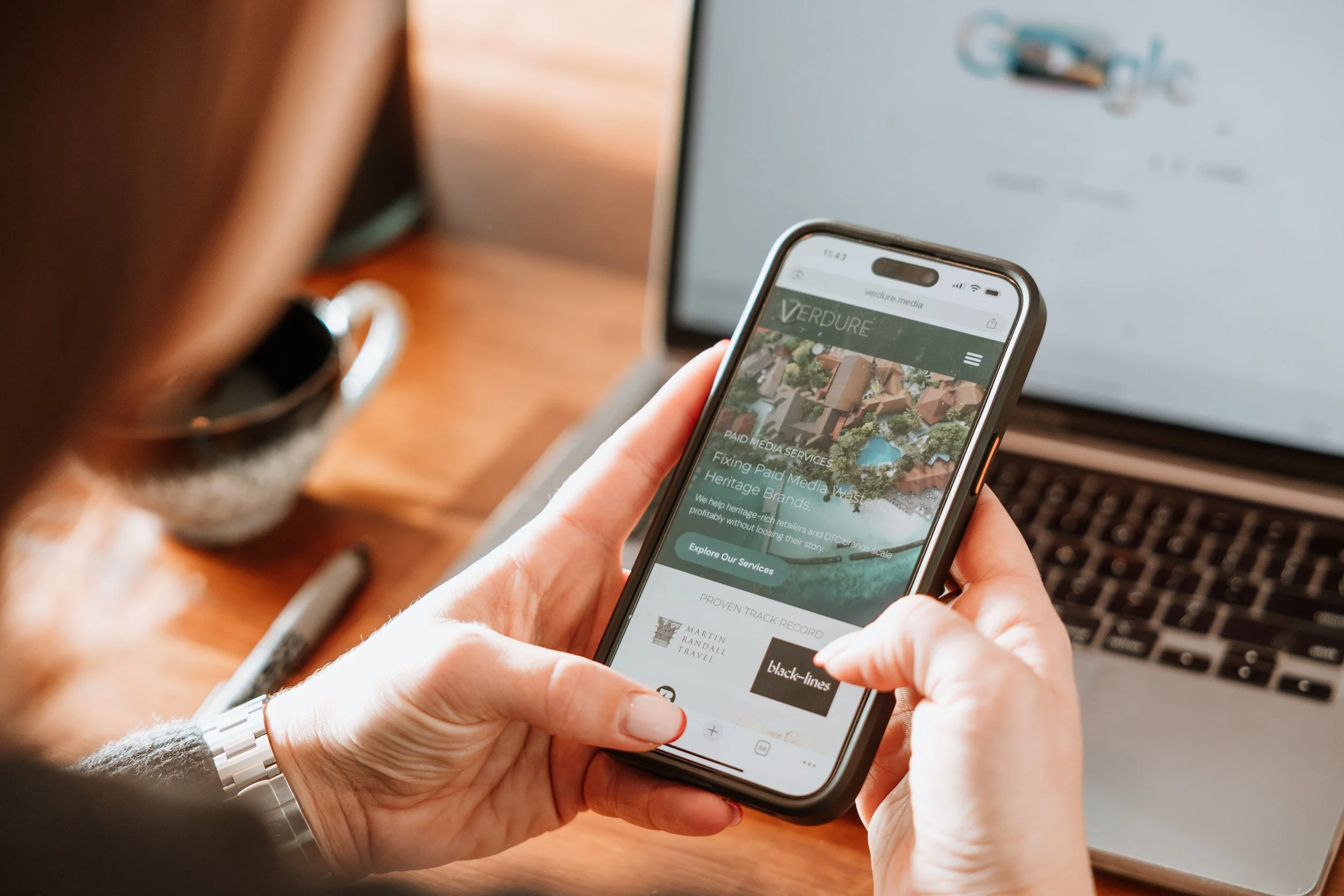 Person holding a smartphone with a website open, sitting at a desk with a laptop and a black pen nearby. The website displayed on the phone is related to media services. Personal Branding by Lucy Rebecca Photography.
