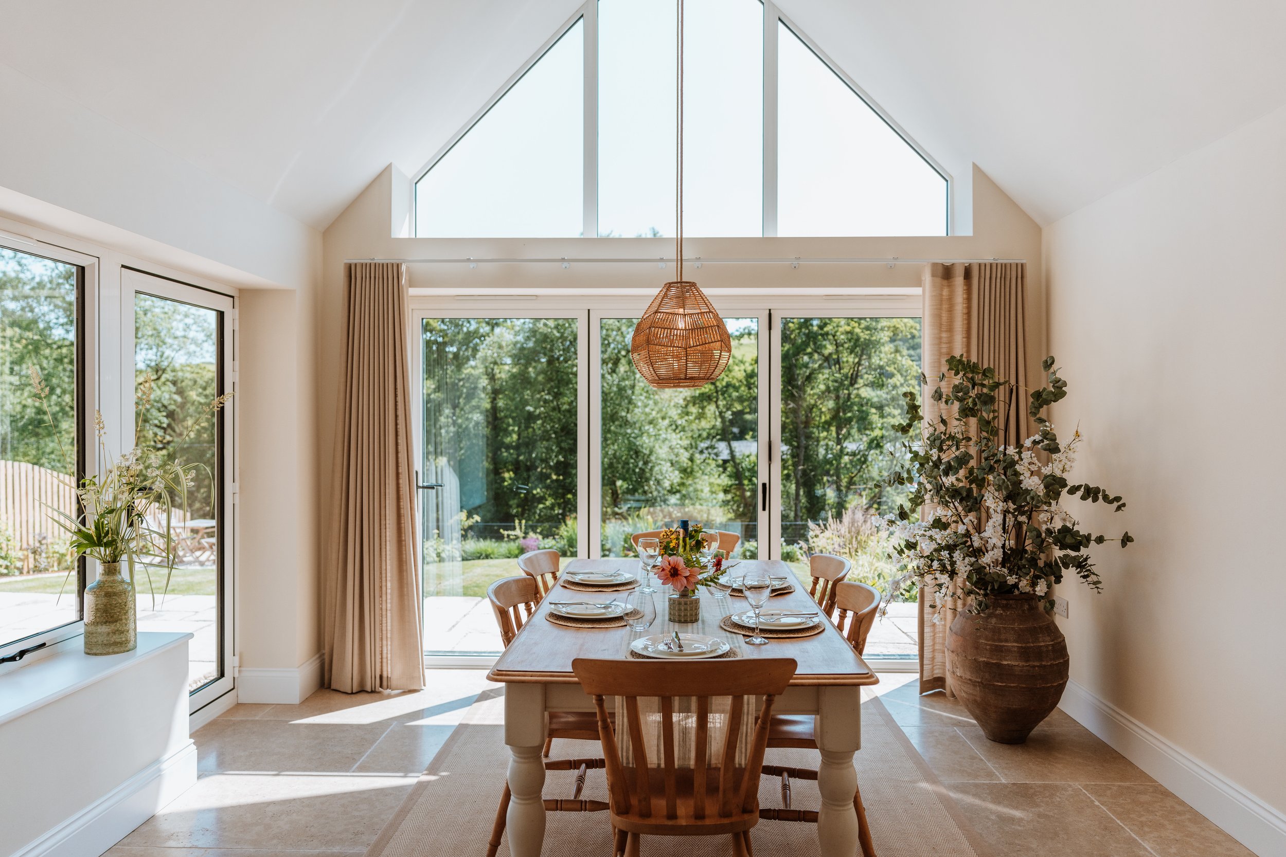 Dining room with a wooden table set with plates, glasses, and a floral centerpiece, large sliding glass doors and windows showing a garden outside, beige curtains, a woven pendant light, and large potted plants. Interiors by Lucy Rebecca Photography.