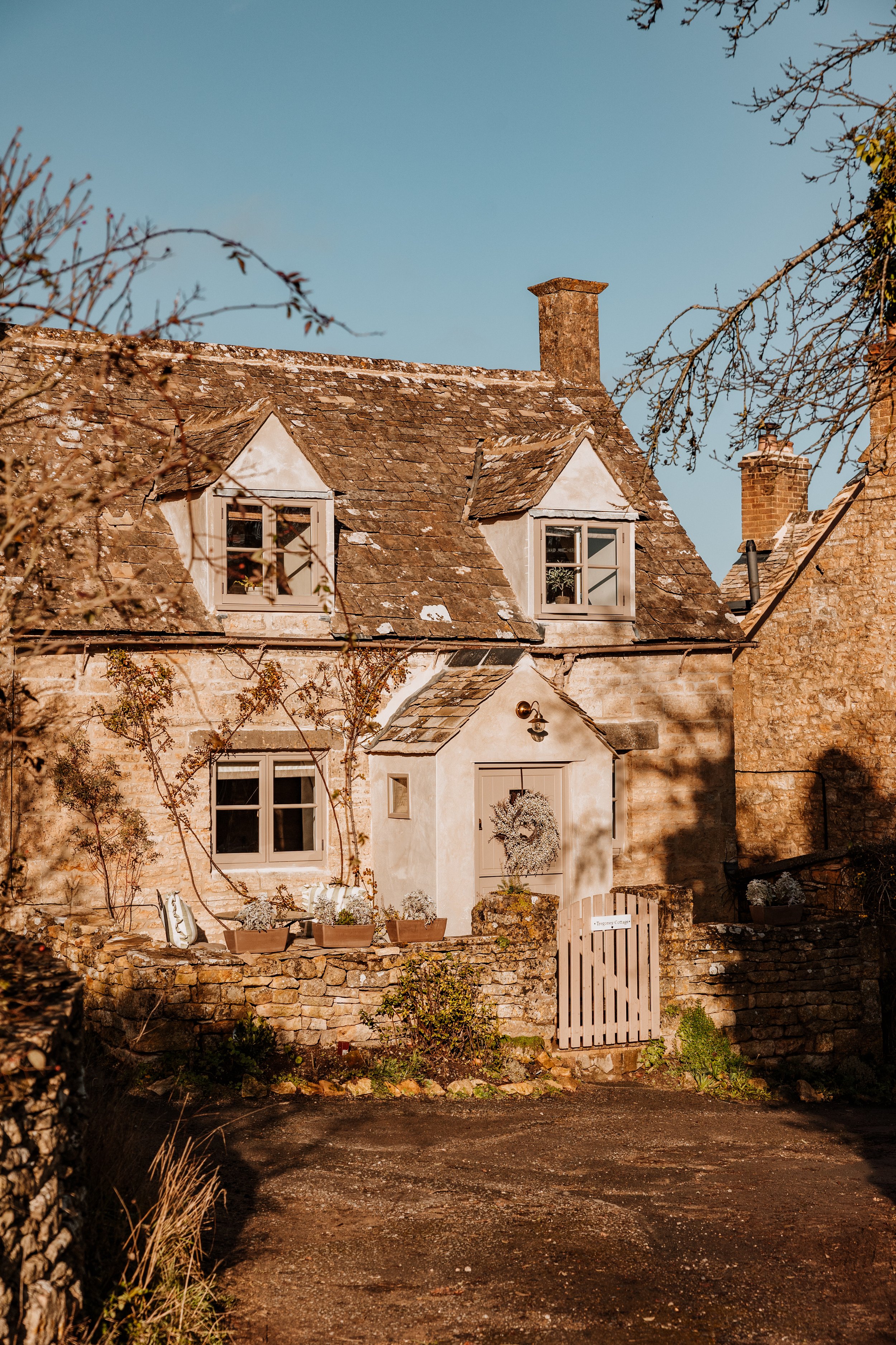 A charming old brick house with a sloped roof, dormer windows, and a small front garden with potted plants, under a clear blue sky. Interiors by Lucy Rebecca Photography.