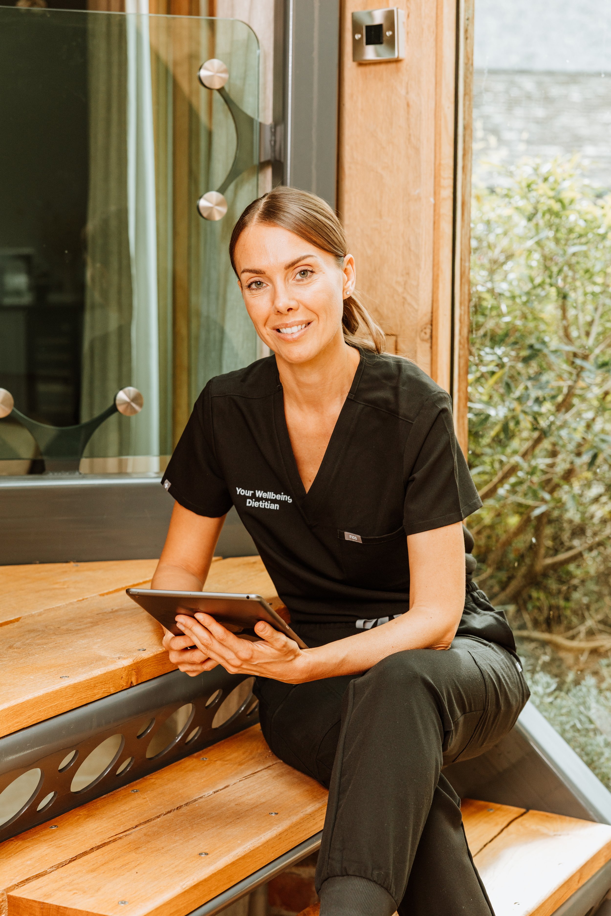 A woman with brown hair tied back, wearing a black uniform with the words "Your Wellbeing Dietitian" on it, sitting on a wooden bench in a bright room with large windows, holding a tablet, and smiling at the camera. Personal Branding by Lucy Rebecca 