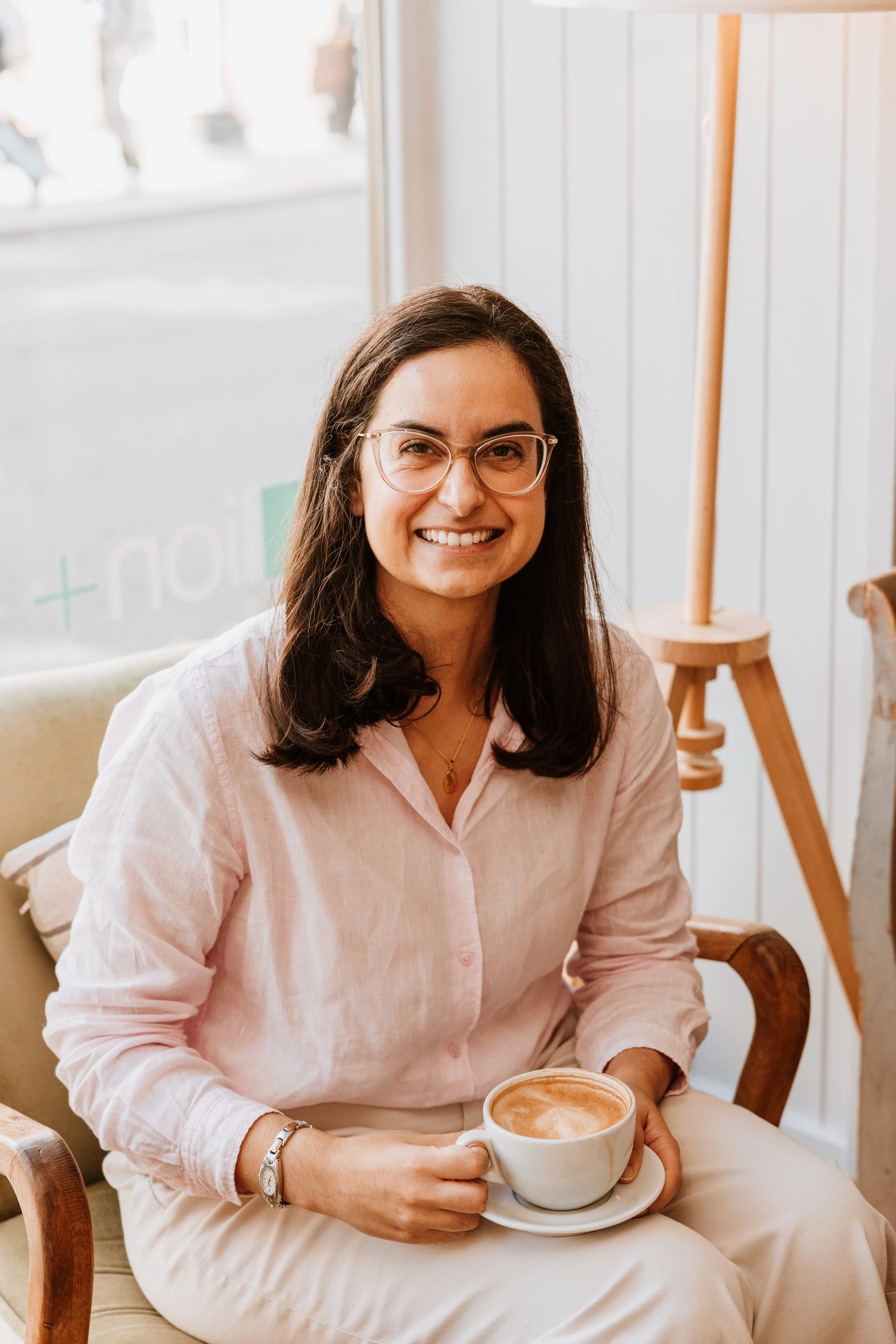 A woman with glasses and dark hair smiling and holding a cup of coffee, sitting in a cafe with light-colored decor. Personal Branding by Lucy Rebecca Photography.