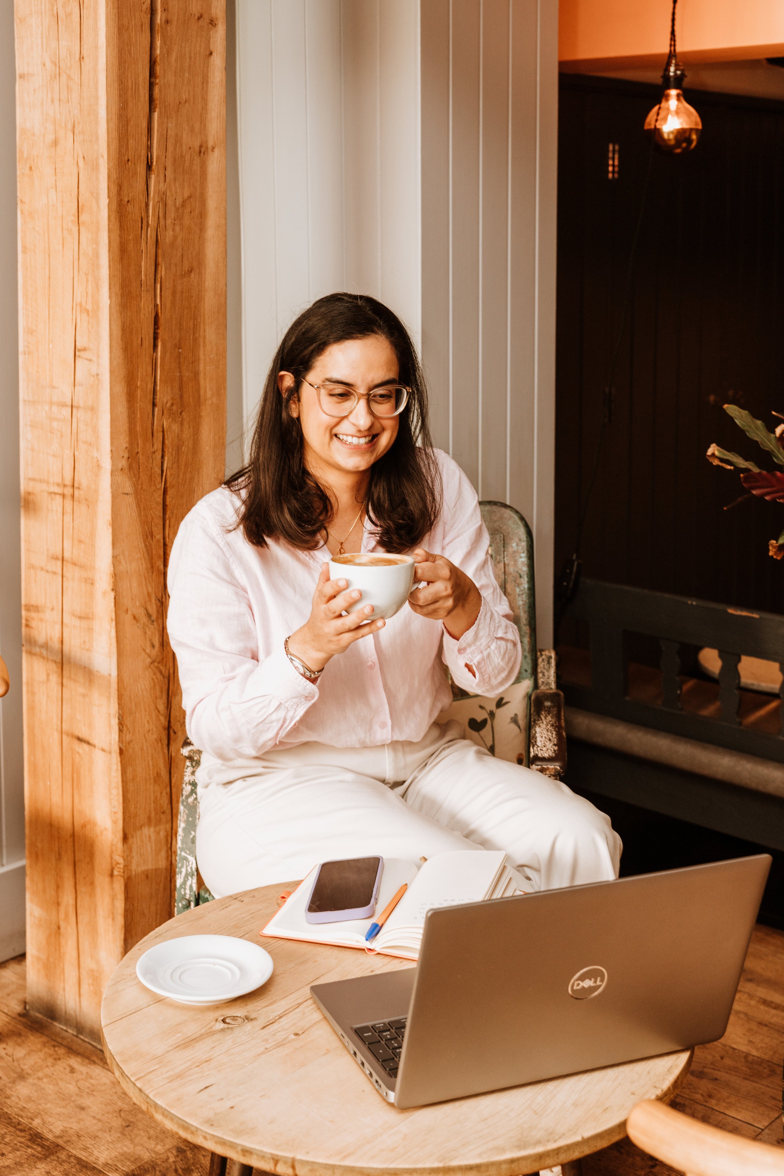 A woman sitting at a wooden table, holding a cup of coffee, smiling, with a laptop, notebook, pen, and smartphone on the table, in a cozy cafe setting. Personal Branding by Lucy Rebecca Photography.