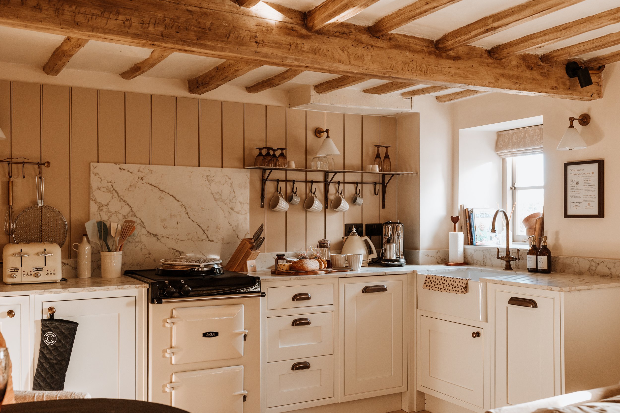 A cozy kitchen with cream-colored cabinets, a vintage stove, marble countertops, and wooden beams on the ceiling. Small kitchen appliances and utensils are on the counters, and cups hang from a wall-mounted rack. A window above the sink lets in natur