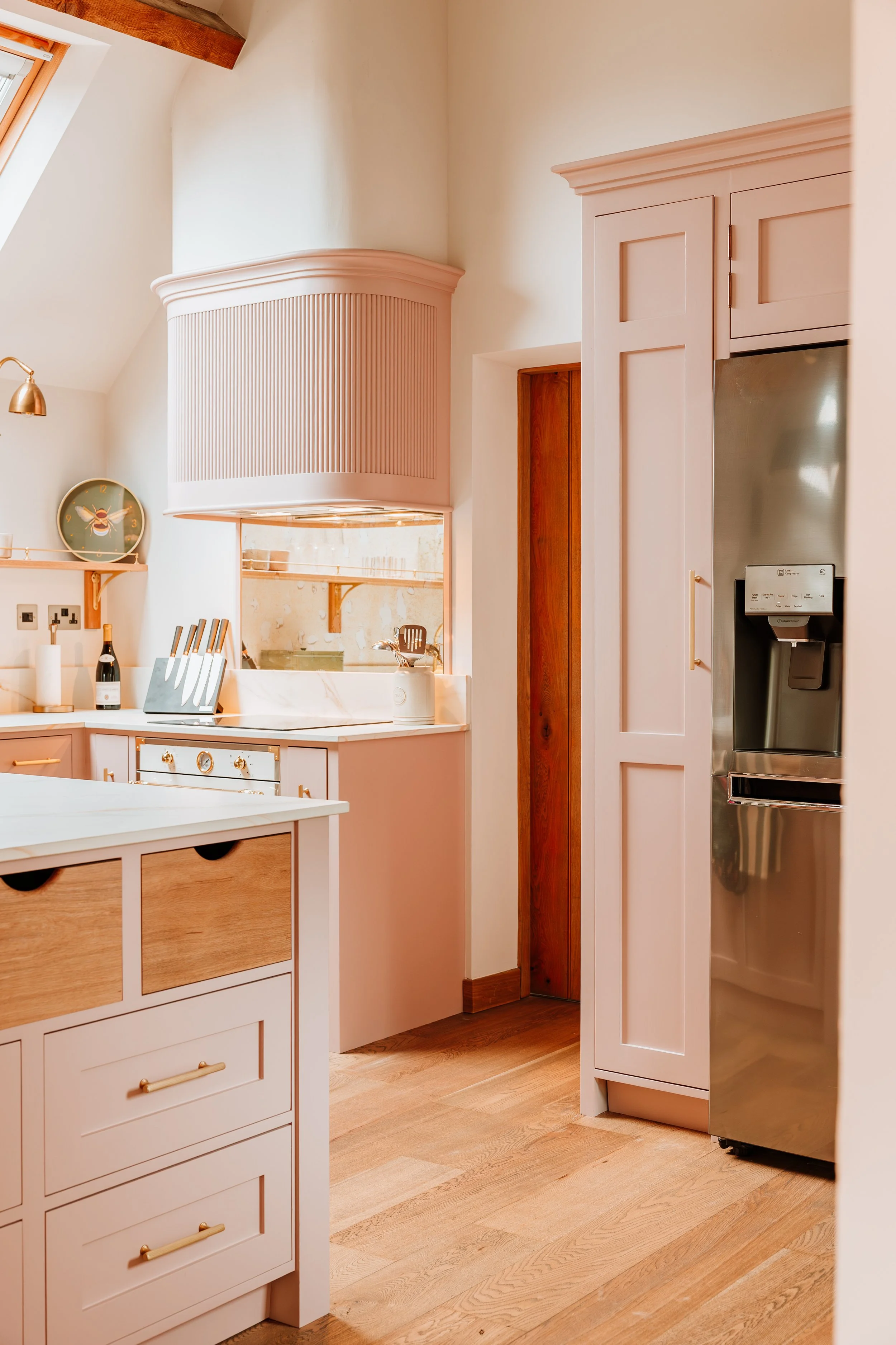 A modern kitchen with white and wood cabinetry, a stainless steel refrigerator, and a wooden floor. There is a small opening to the outside or another room with a counter and some kitchen utensils. Interiors by Lucy Rebecca Photography.