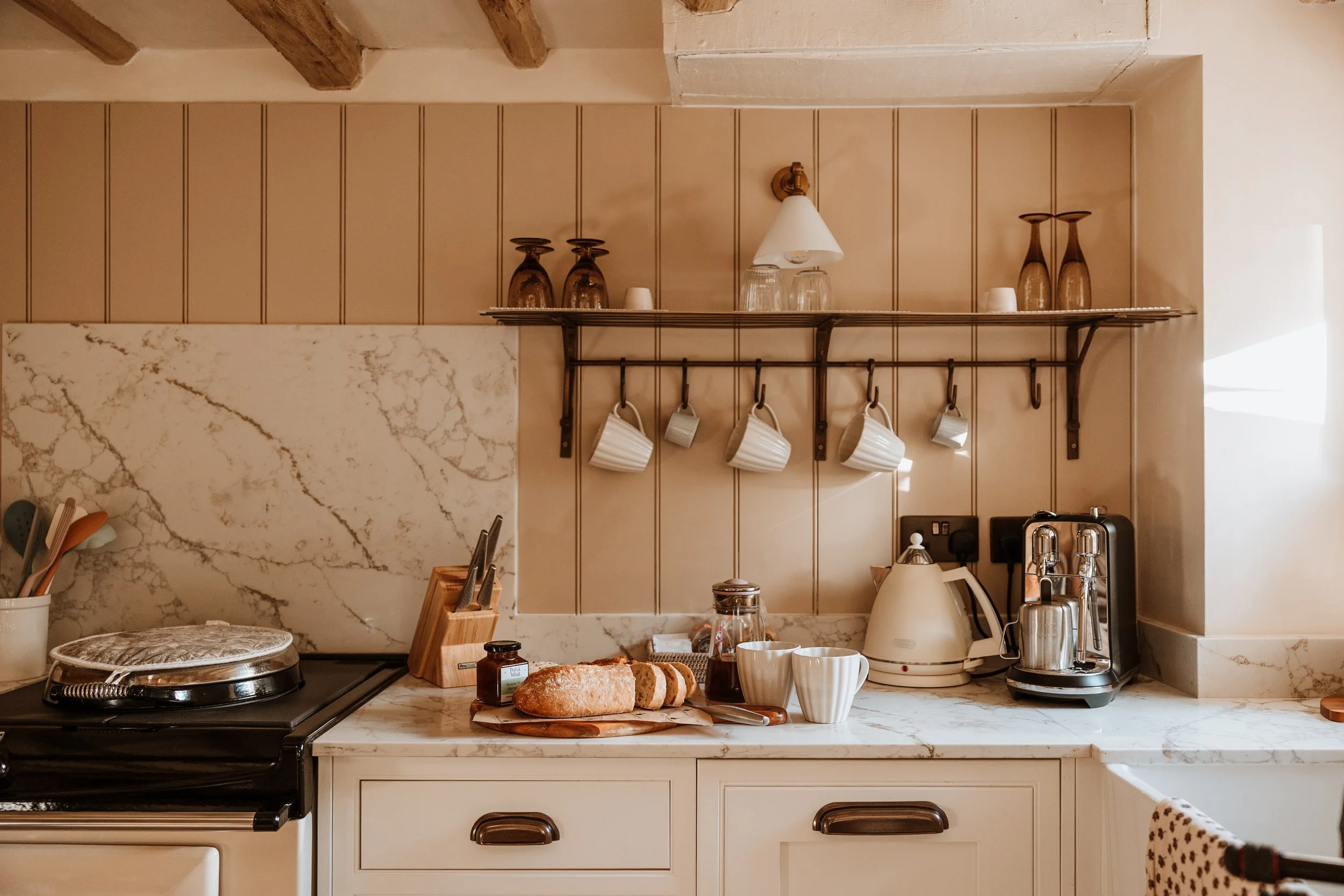 Kitchen counter with bread, cups, and jars, with hanging mugs and glasses on a shelf above. Interiors by Lucy Rebecca Photography.