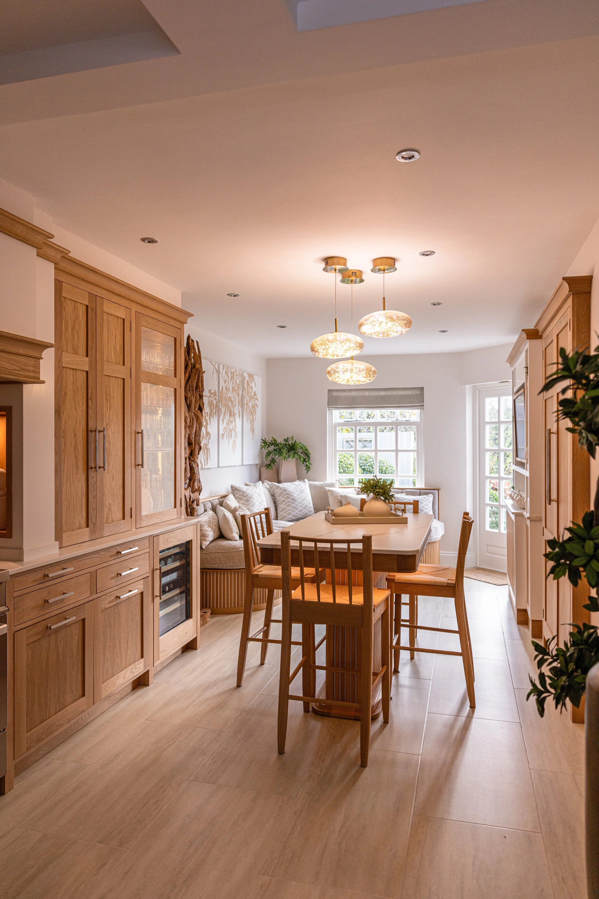 Bright kitchen dining area with wooden cabinets, a central island, a cozy built-in corner nook with pillows, and modern pendant lighting. Interiors by Lucy Rebecca Photography.