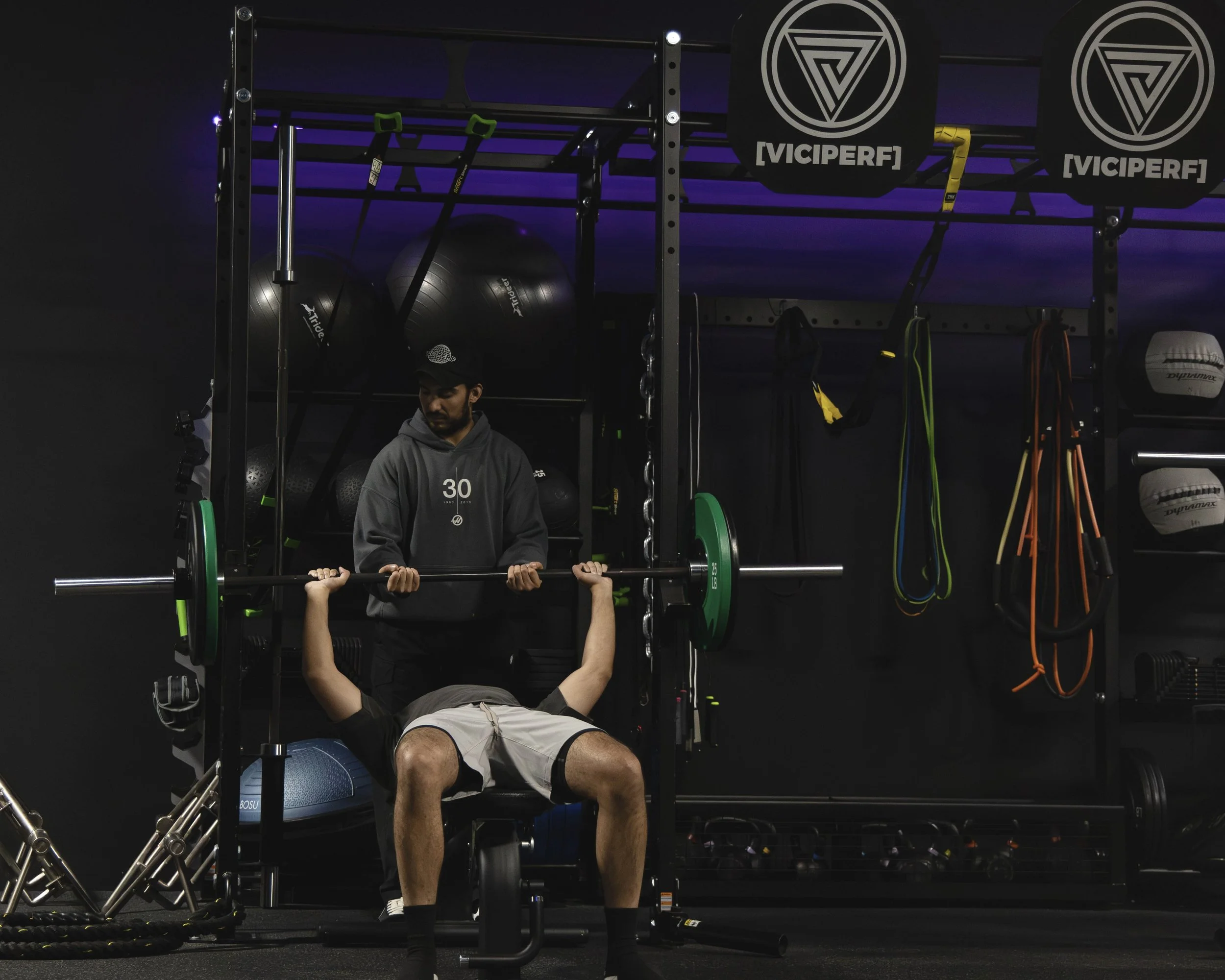 Man bench pressing with weights in a gym, with a trainer standing behind him, surrounded by fitness equipment and colorful resistance bands hanging on a black wall.