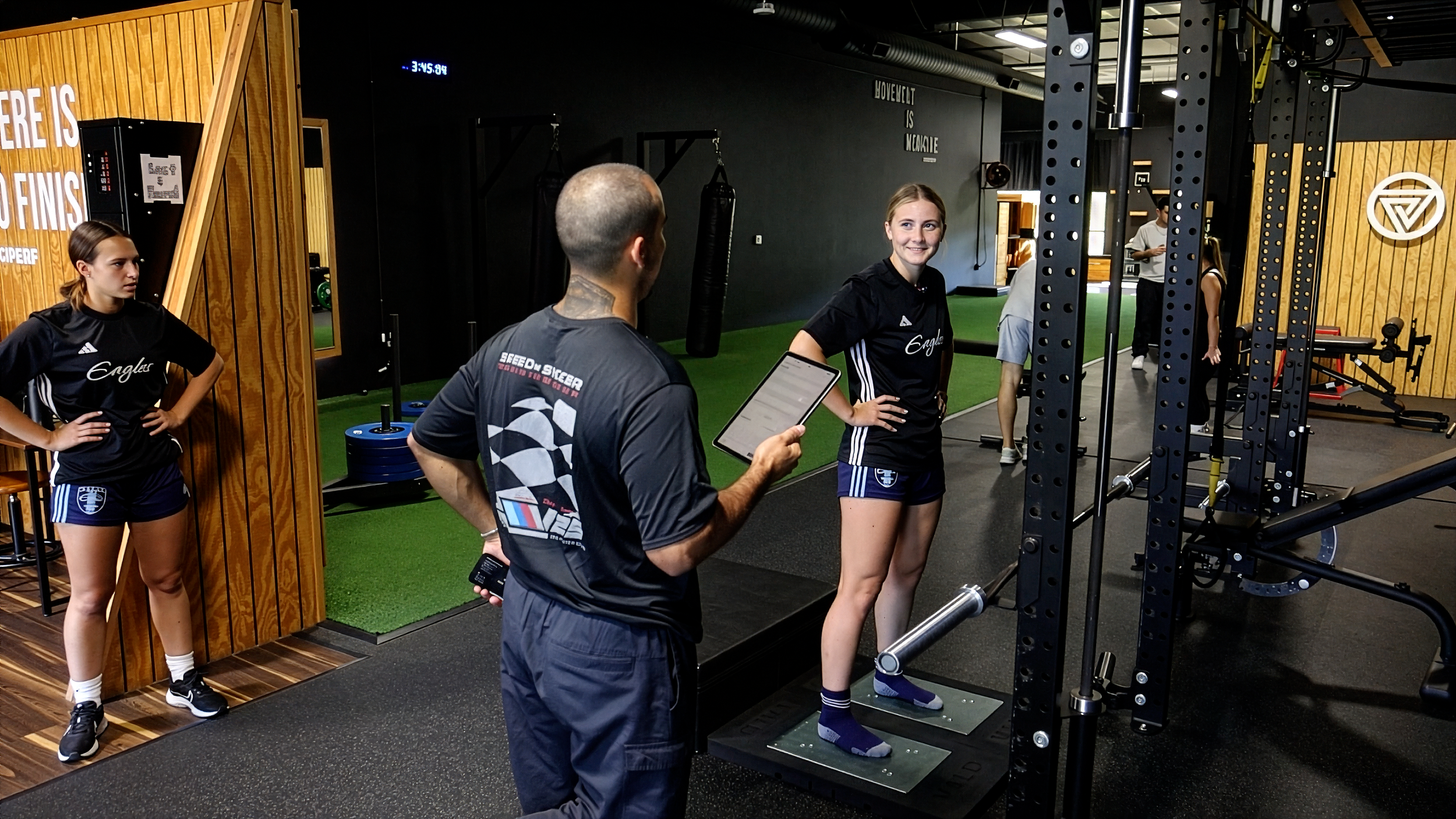 A young woman standing on an indoor athletic scale, being monitored by a man holding a tablet, inside a gym with other people in the background.
