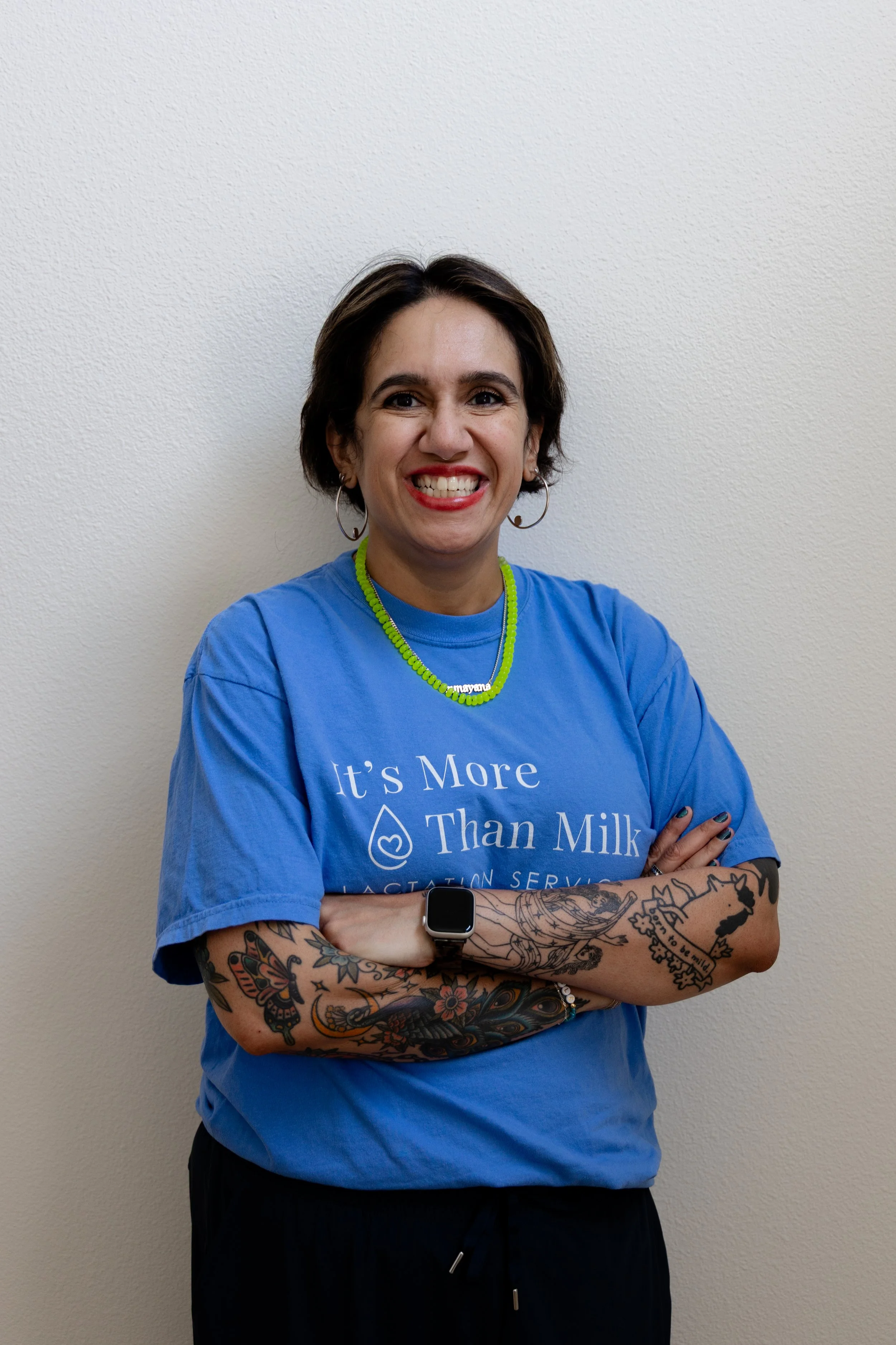 A woman with curly dark hair wearing a black T-shirt with the words 'Inform. Support. Empower.' printed on it, standing against a pink wall. She has jewelry including a green beaded necklace, gold hoop earrings, and a silver wristwatch, and has tattoos on her left arm and hand.