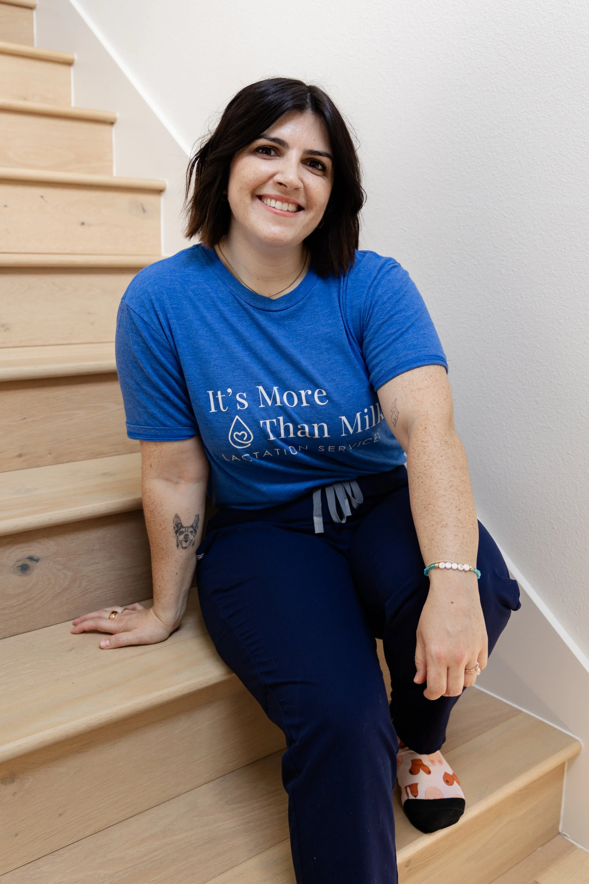 A woman with shoulder-length dark hair, wearing a black T-shirt with white text, and light blue jeans, standing in front of a pink wall, smiling and posing with one hand in her pocket.