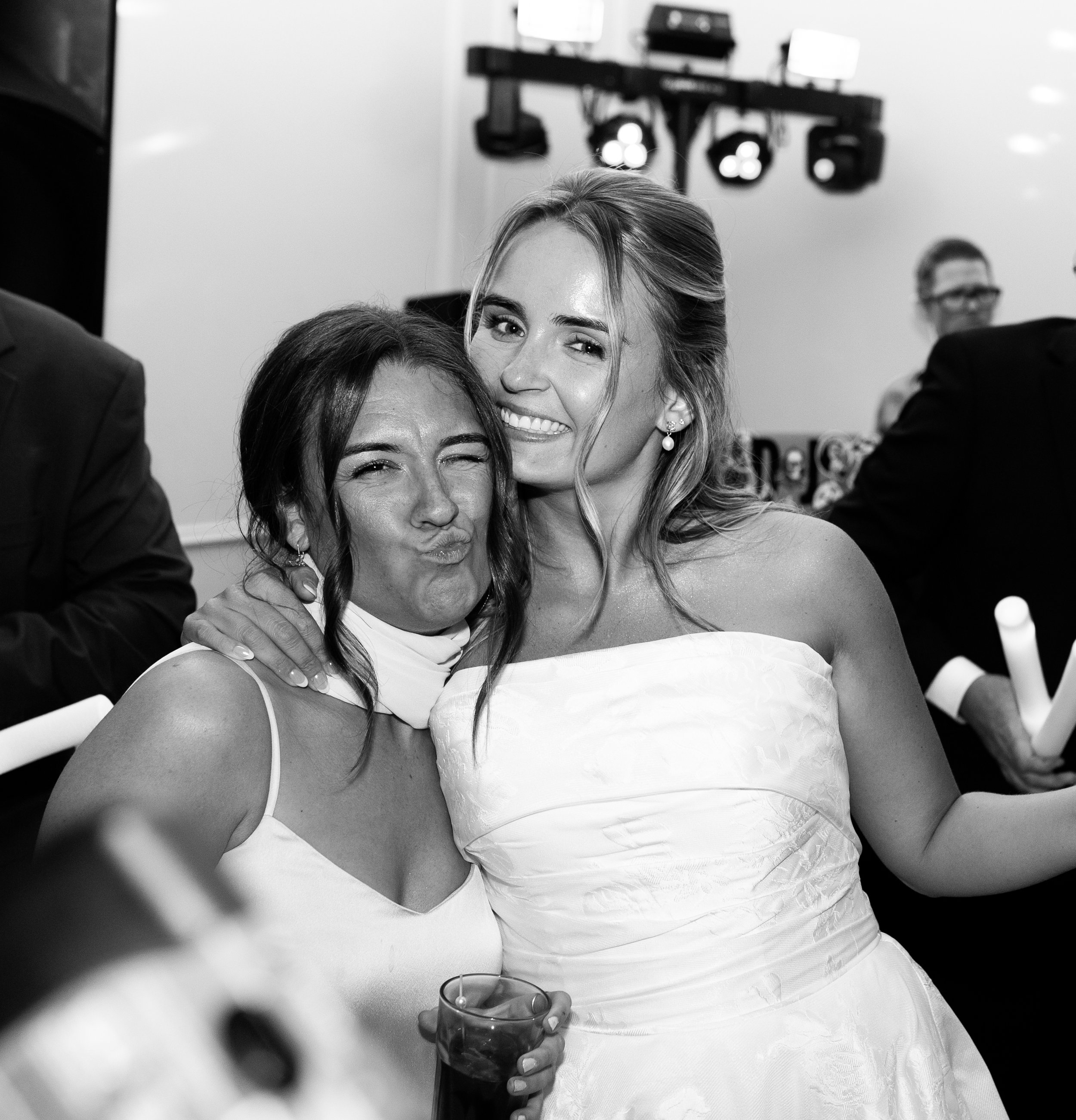 Two women celebrating at a wedding reception, one in a white dress and the other in a sleeveless top, smiling and hugging each other, with a dance floor and lights in the background.