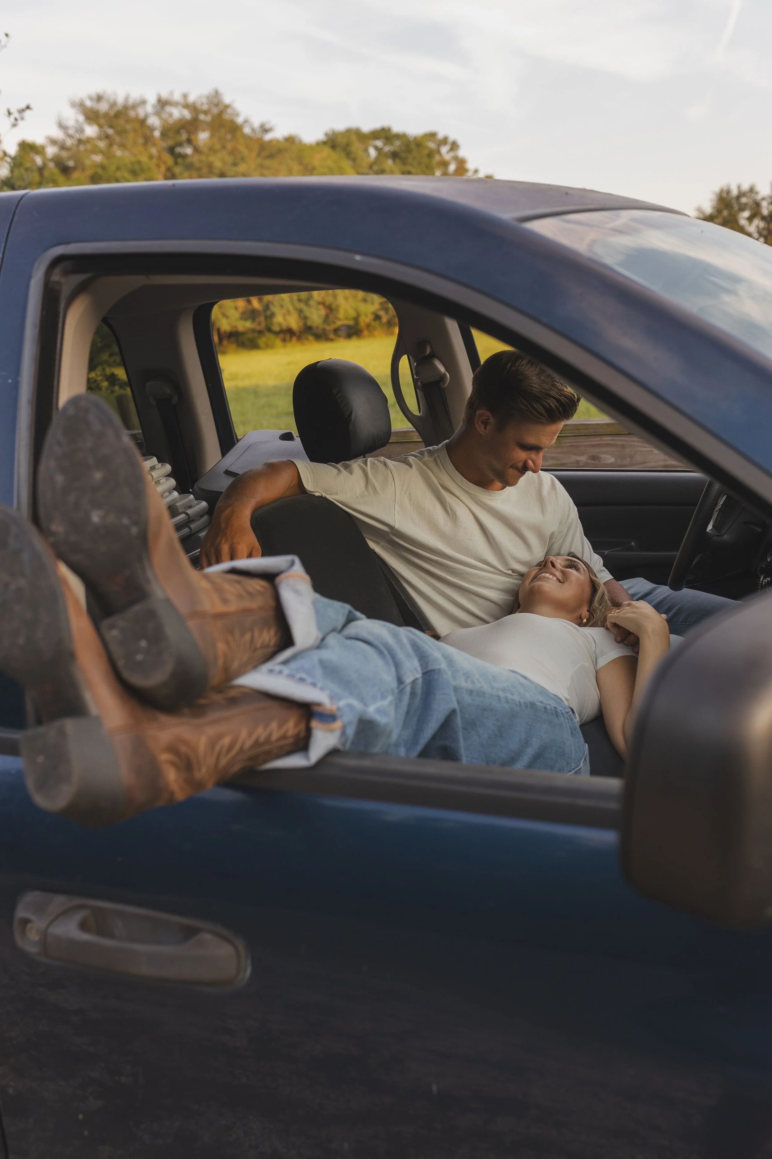 A man sitting in a pickup truck with his feet up on the door, a woman lying across the front seats, smiling and looking at him, with a grassy field and trees in the background during daylight.