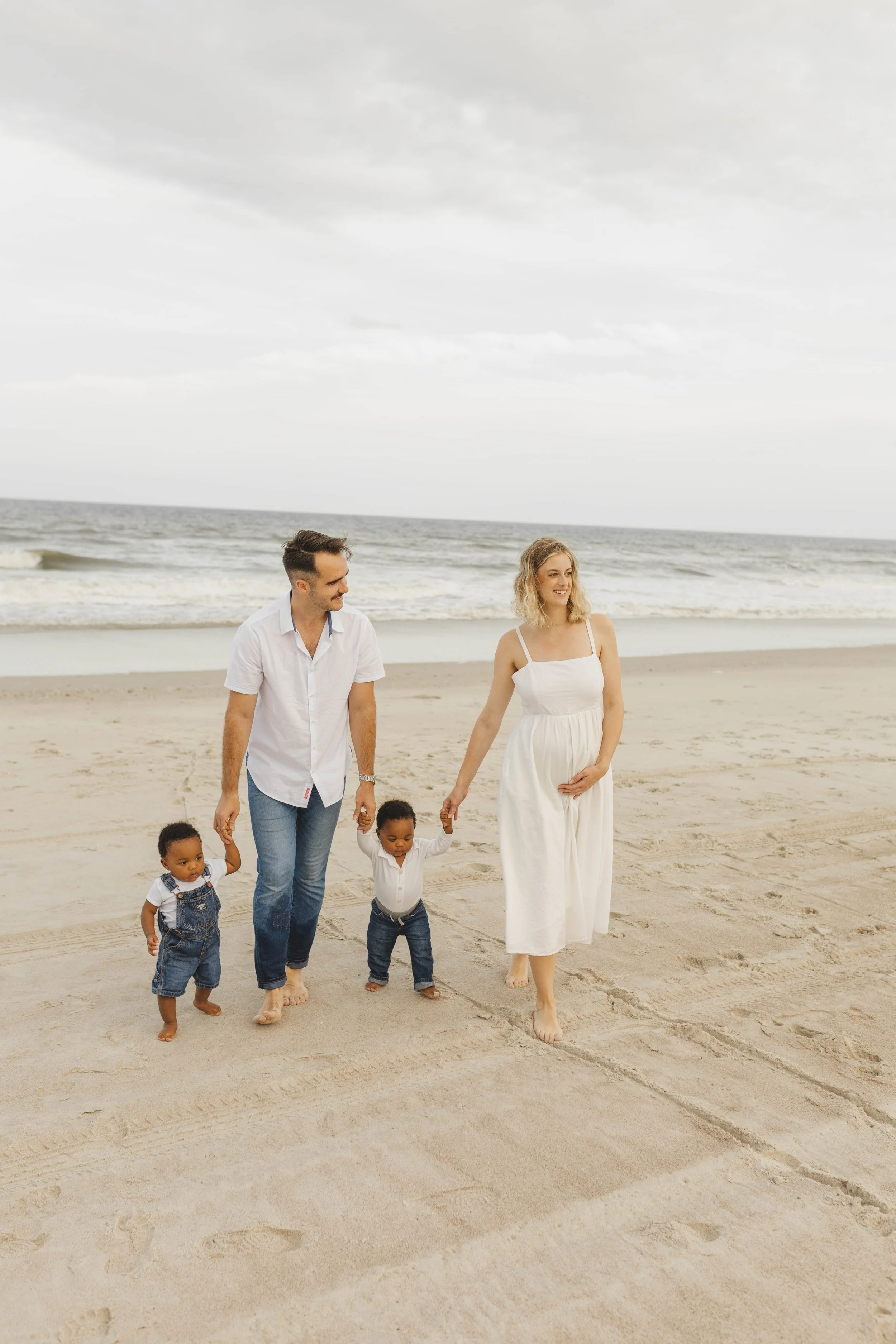 A family of four walking on the beach, holding hands, with the ocean in the background. The woman is pregnant, wearing a white dress. The man and two children are dressed casually, with the children in denim overalls and white shirts.