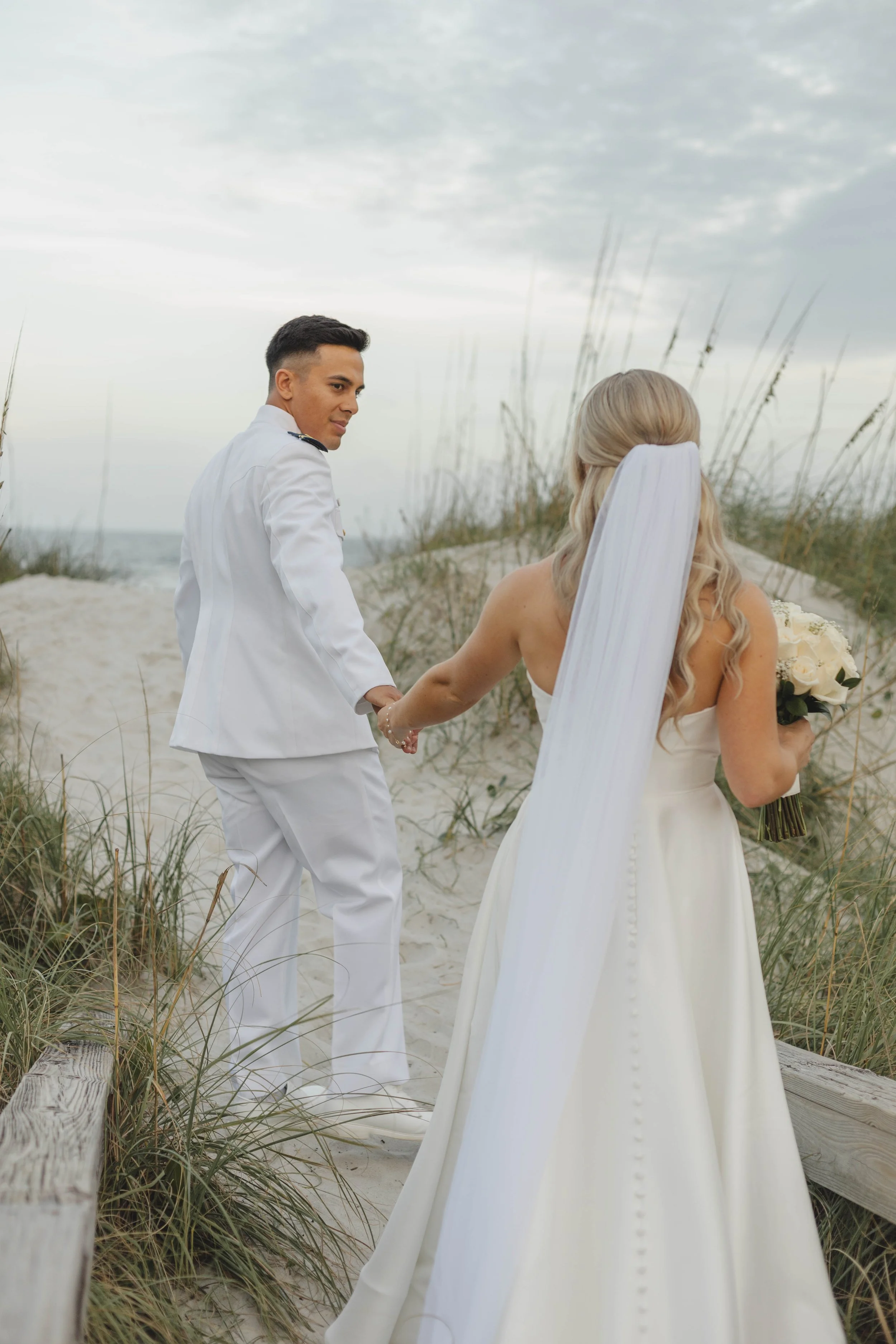 A bride and groom holding hands on a beach, with the groom in a white sailor suit and the bride in a white wedding gown holding a bouquet, as waves and sky are in the background.