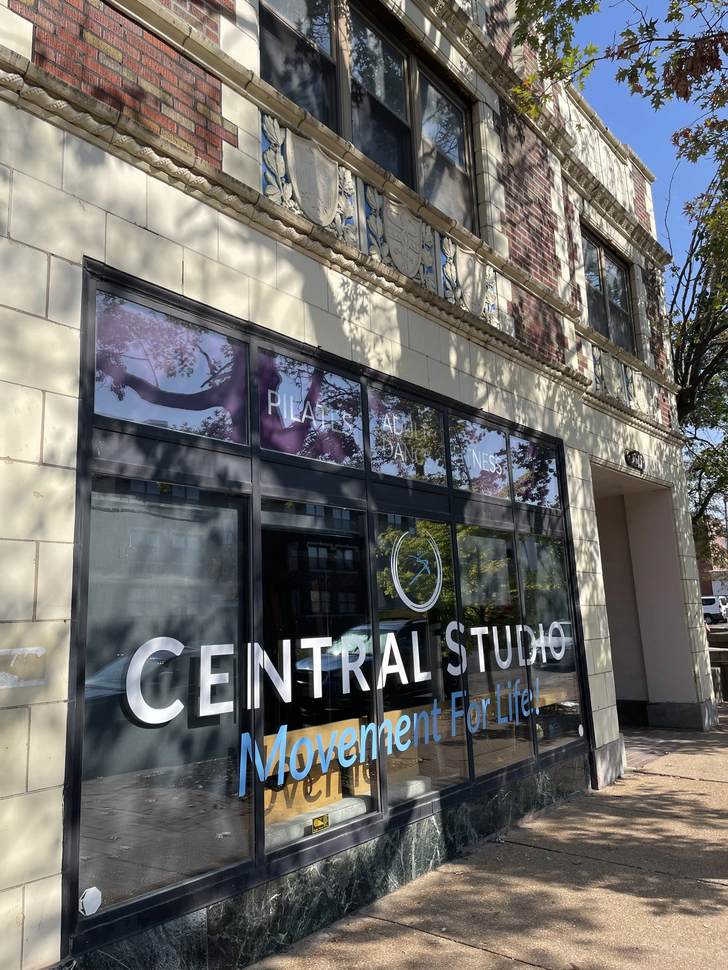 Storefront with large glass windows displaying the words "Central Studio Movement For Life!" and a stylized clock logo. The building has decorative architectural details and is partially shaded by nearby trees.