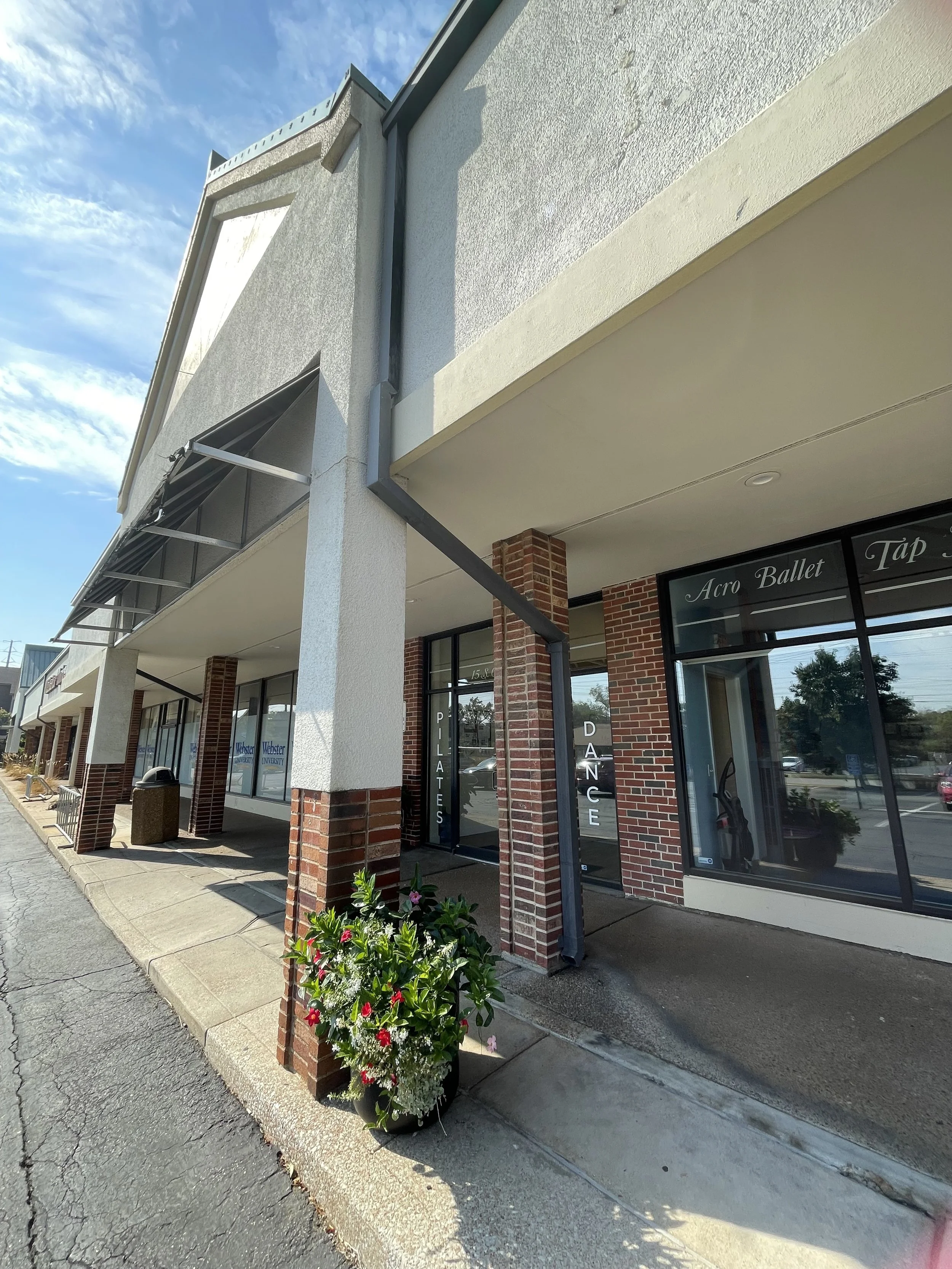 Exterior view of a commercial building with brick columns, large glass windows, and a flower pot with pink and white flowers near the sidewalk. Signs indicate dance and ballet classes inside.
