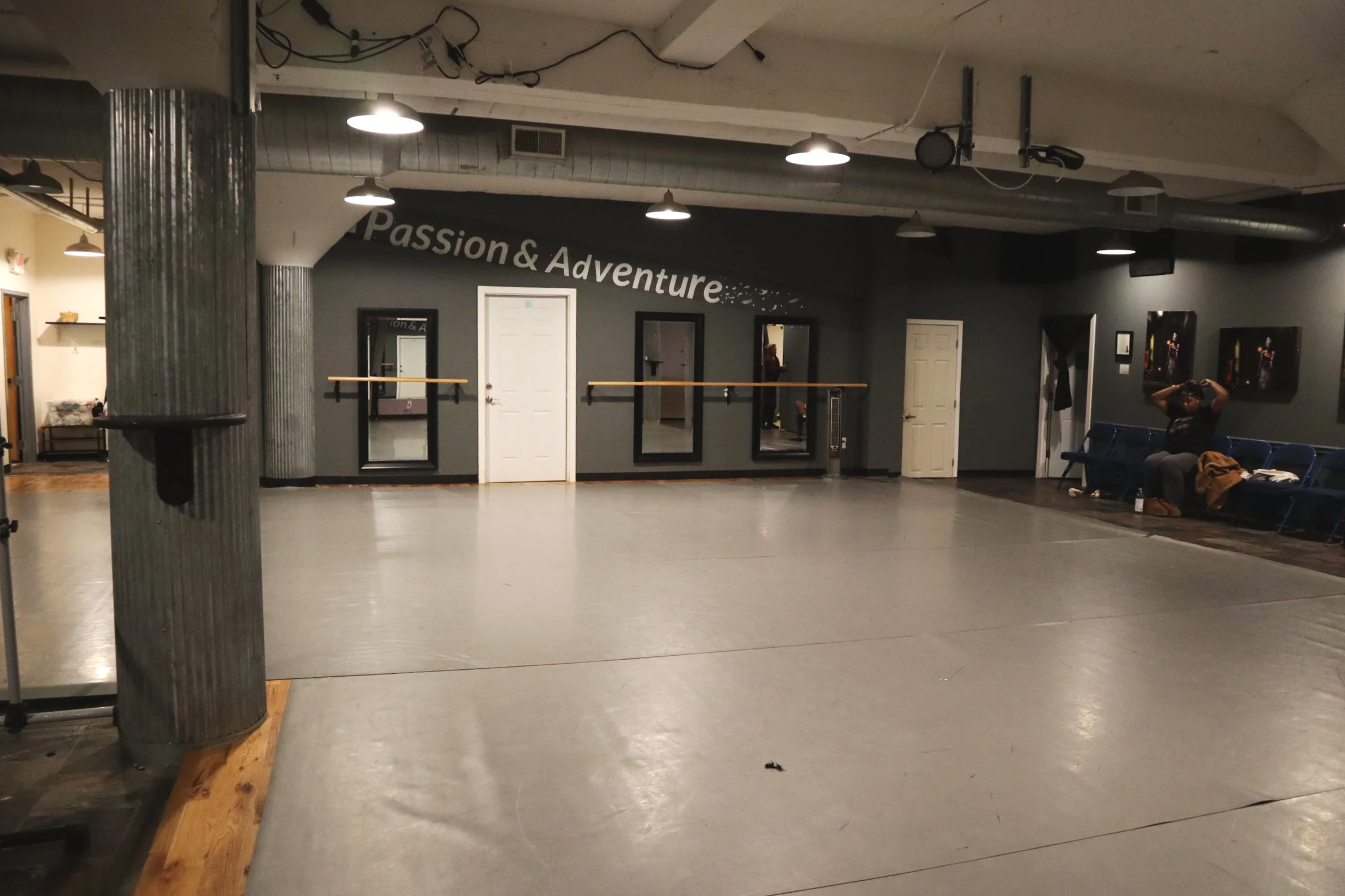 An empty dance studio with a large open floor, wall-mounted mirrors, ballet barres, and a wall sign that reads "Passion & Adventure." There are some chairs with a person sitting and taking photos, and a few framed pictures on the wall.