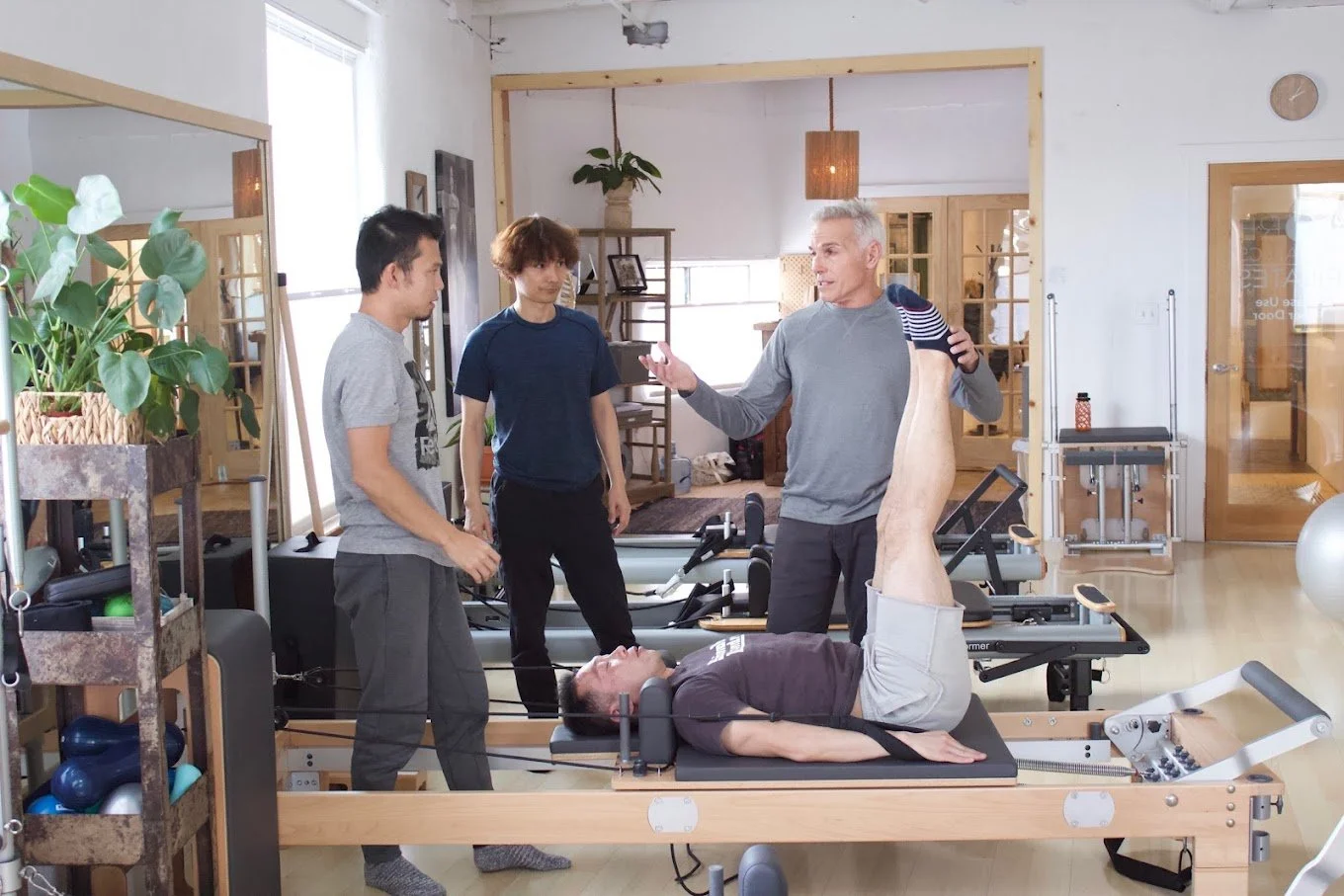 Four individuals in a Pilates studio, one lying on a reformer with legs extended, others observing and discussing.