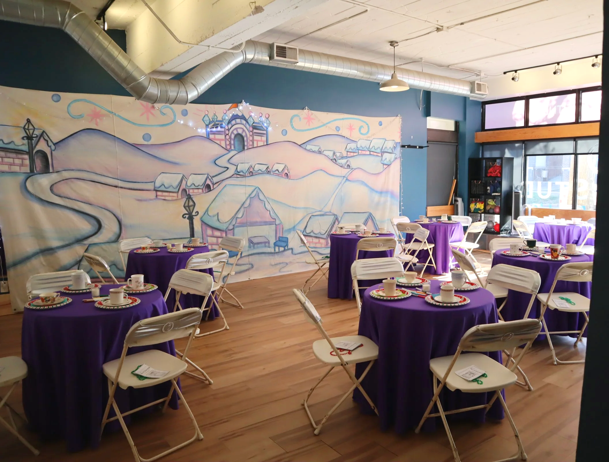 A decorated banquet room with purple tablecloths, white folding chairs, and a mural of a winter village scene with snow-covered houses and a castle on a blue wall.