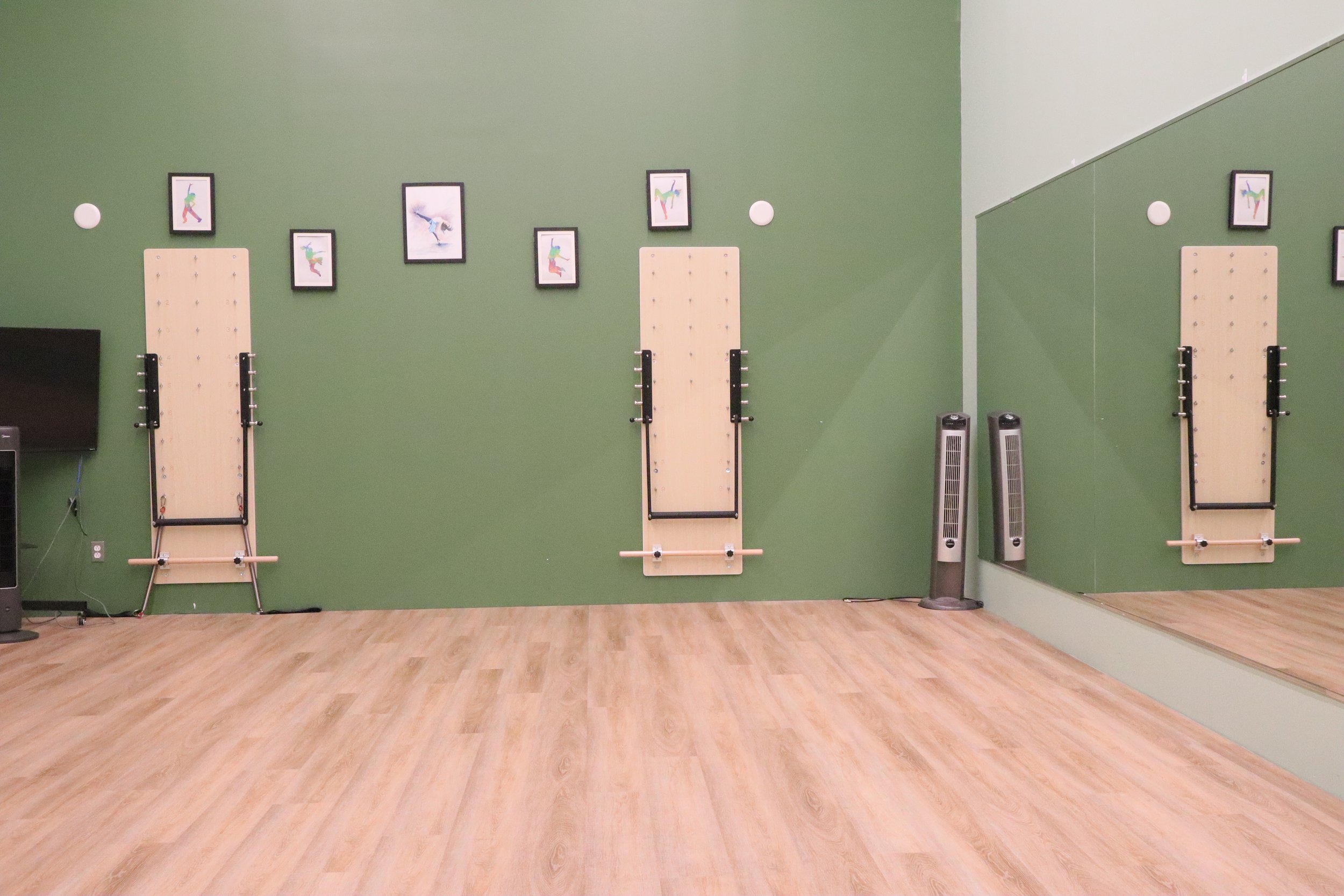 A dance or exercise studio with a green wall, a large mirror on the right, wooden floors, and three wall-mounted wooden dance boards with black handrails. There are framed pictures of dancers above the boards, two white circular wall decorations, and two tall electric fans on the right side.
