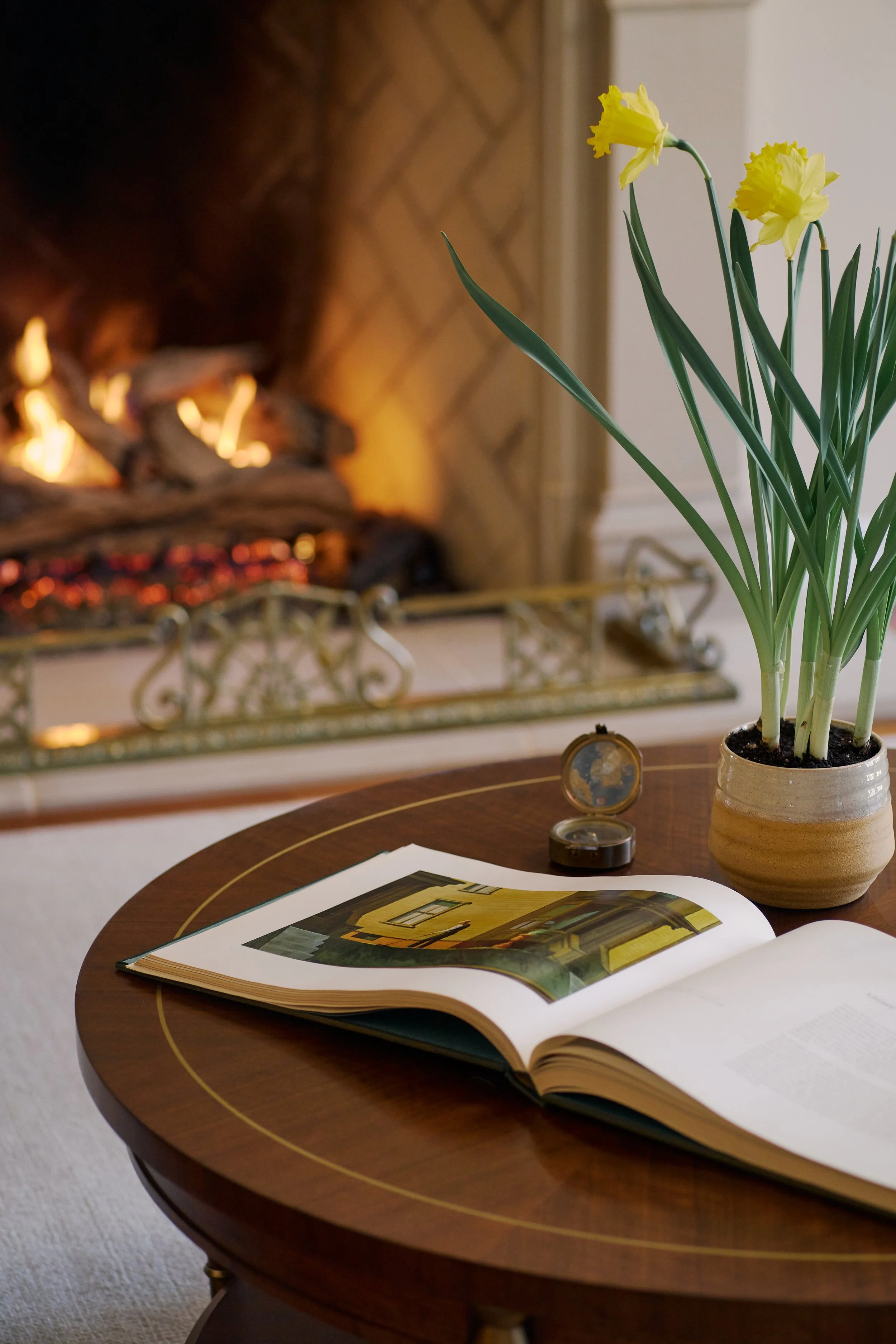 A round wooden table with an open book, a small clock, and a potted yellow daffodil plant in front of a fireplace with a fired burning log inside.