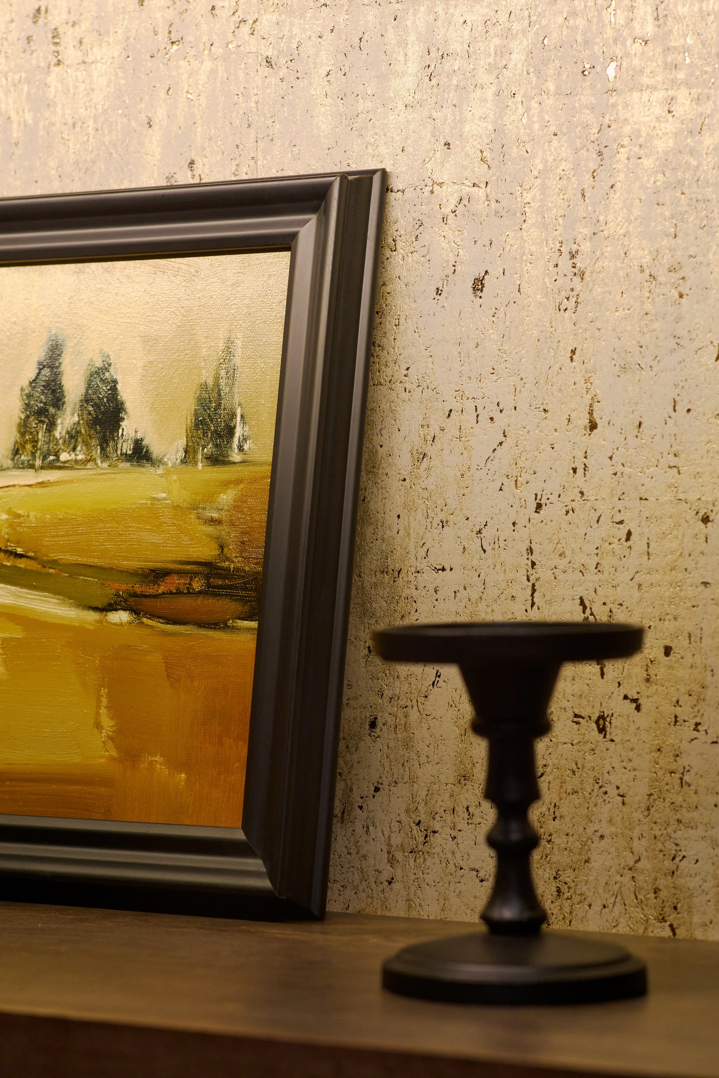 Close-up of a framed painting of a landscape with trees, resting on a wooden surface next to a black candle holder against a textured beige wall.