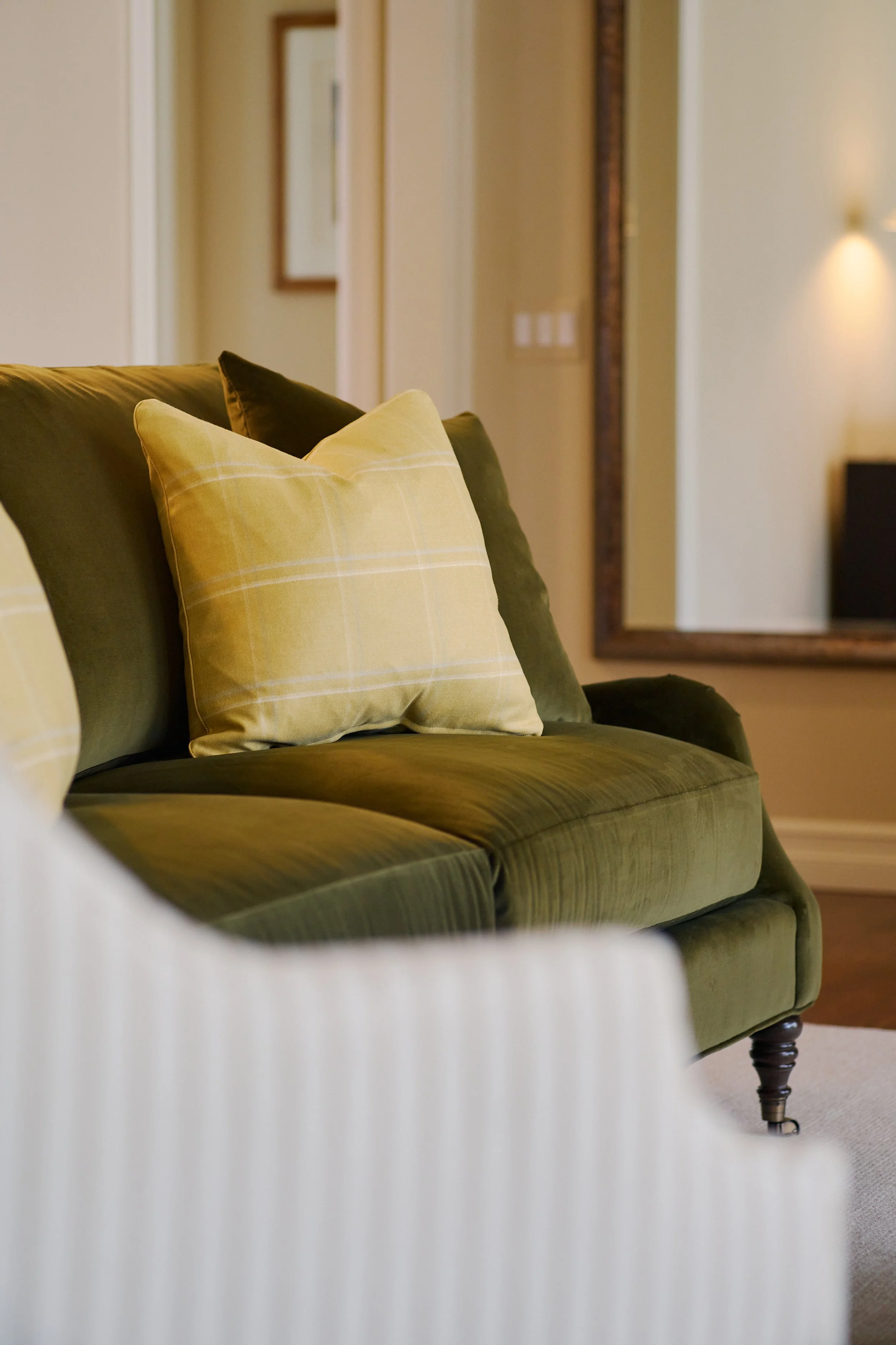 Interior of a living room with a green velvet sofa and yellow plaid cushions, a large wall mirror, and a framed picture in the background.