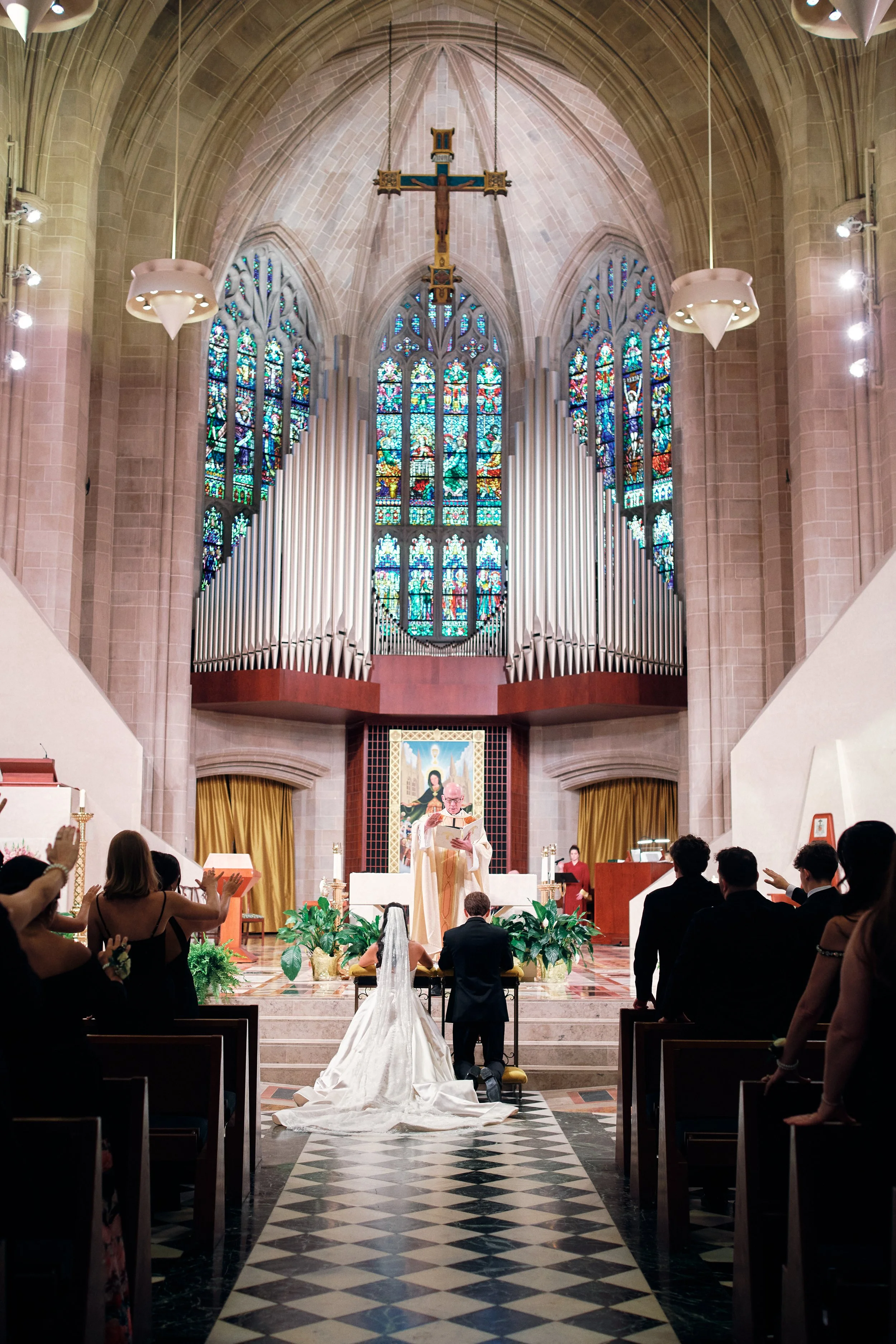 A wedding ceremony taking place inside a grand, ornate church with many guests seated and a couple standing at the altar. At the Cathedral of the Most Blessed Sacrament in Detroit, Michigan.