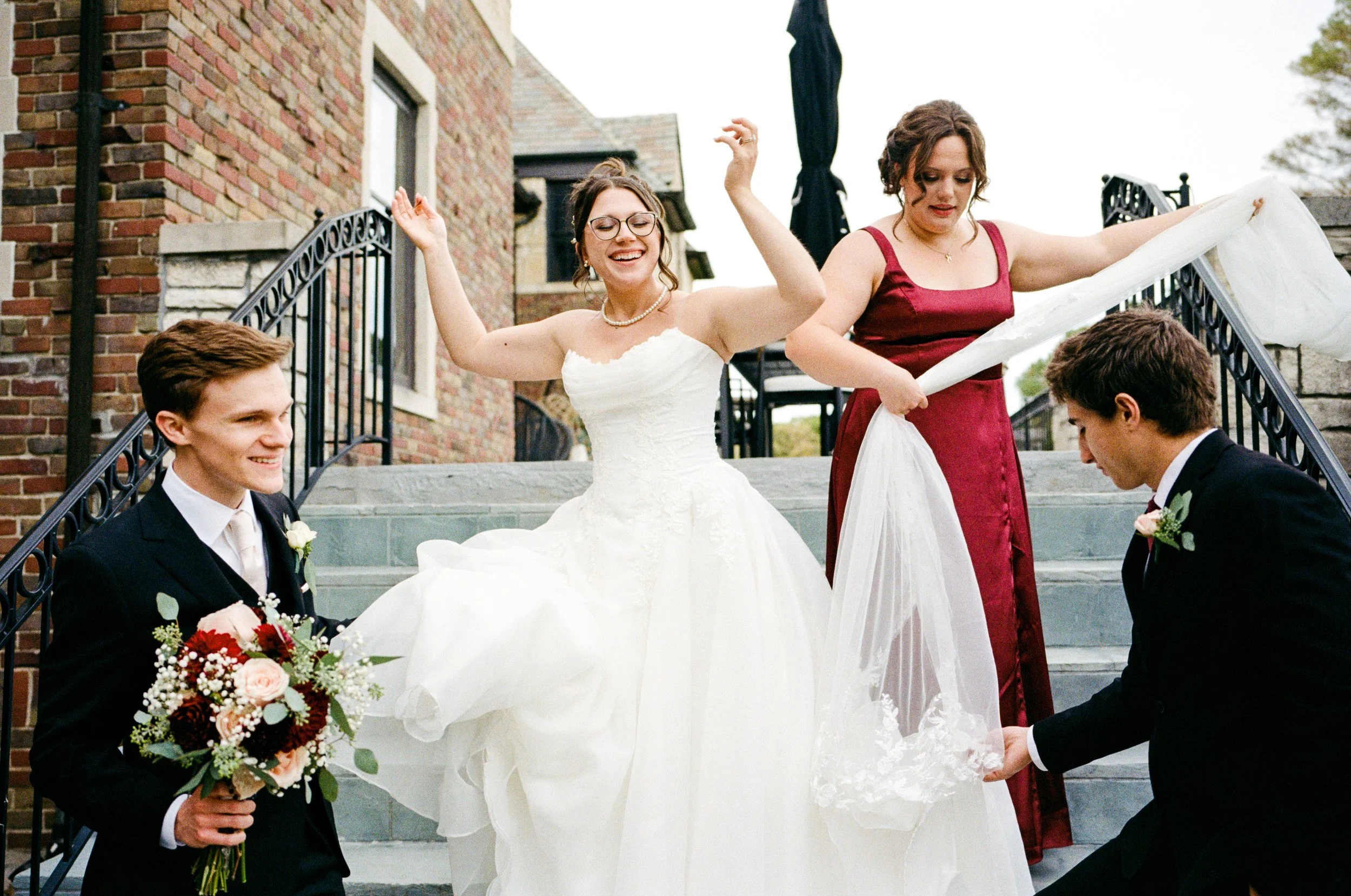 A bride in a white wedding dress happy and smiling, standing on outdoor stairs, being assisted by two women, with two men in suits holding flowers.