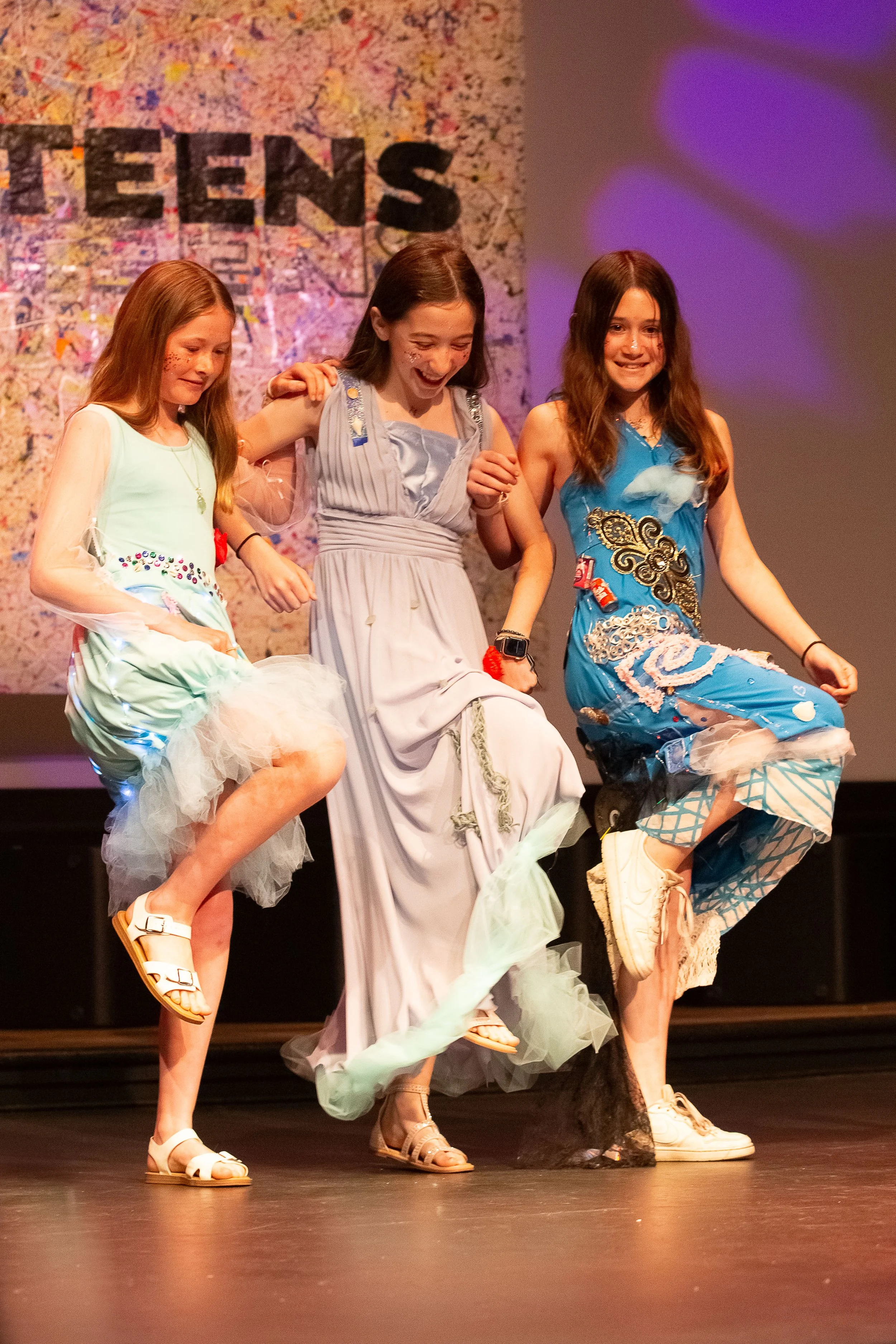 Three FashionTeens Santa Cruz student designers joyfully kick up their heels on the runway in handmade upcycled dresses embellished with tulle, recycled materials, and creative sparkle—showcasing fun, friendship, and sustainable fashion in motion.