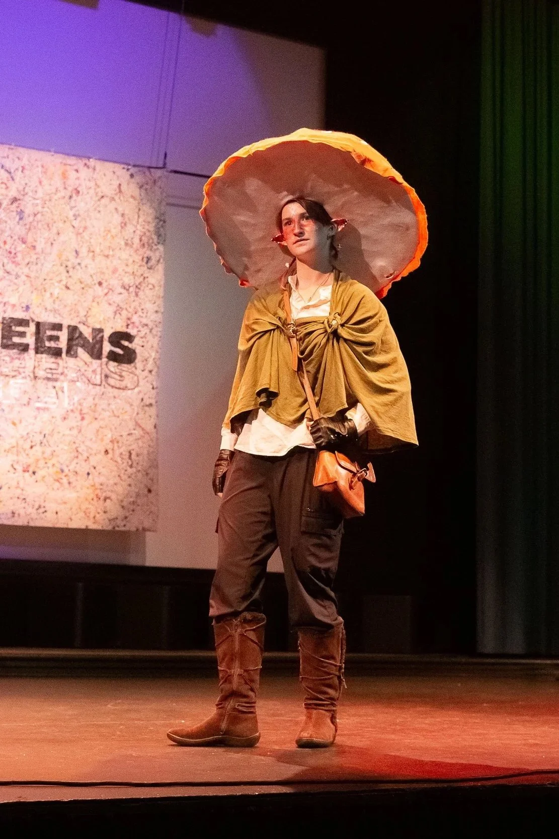 FashionTeens Santa Cruz student model dressed in a whimsical, mushroom-inspired look featuring a giant handmade mushroom cap hat, forest-toned cape, and boots — channeling nature, storytelling, and imaginative upcycled design.