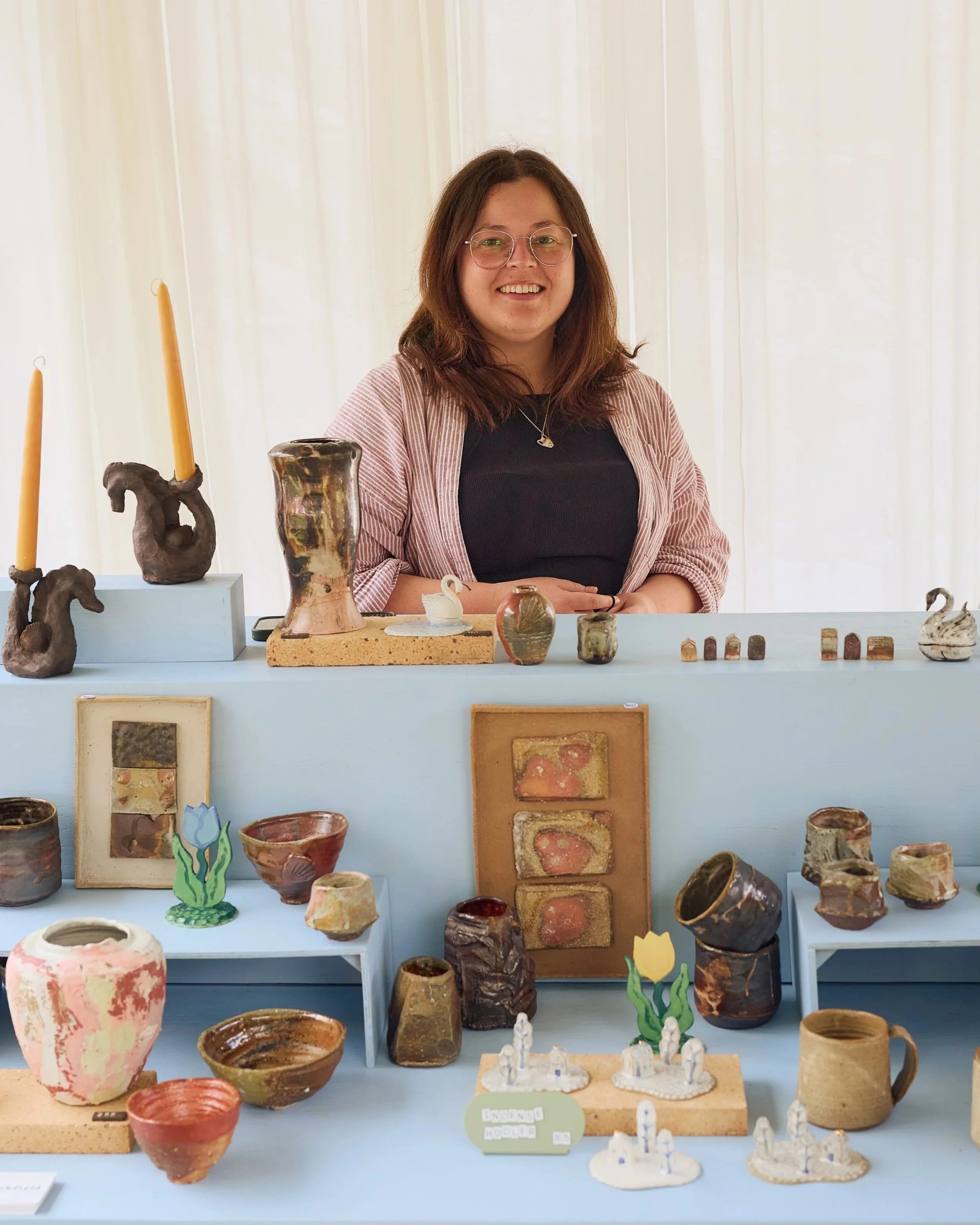 Display of various handmade pottery and ceramic art pieces on a light blue table and shelves, including vases, bowls, and decorative objects at the 25th annual Pottery Expo 2026.