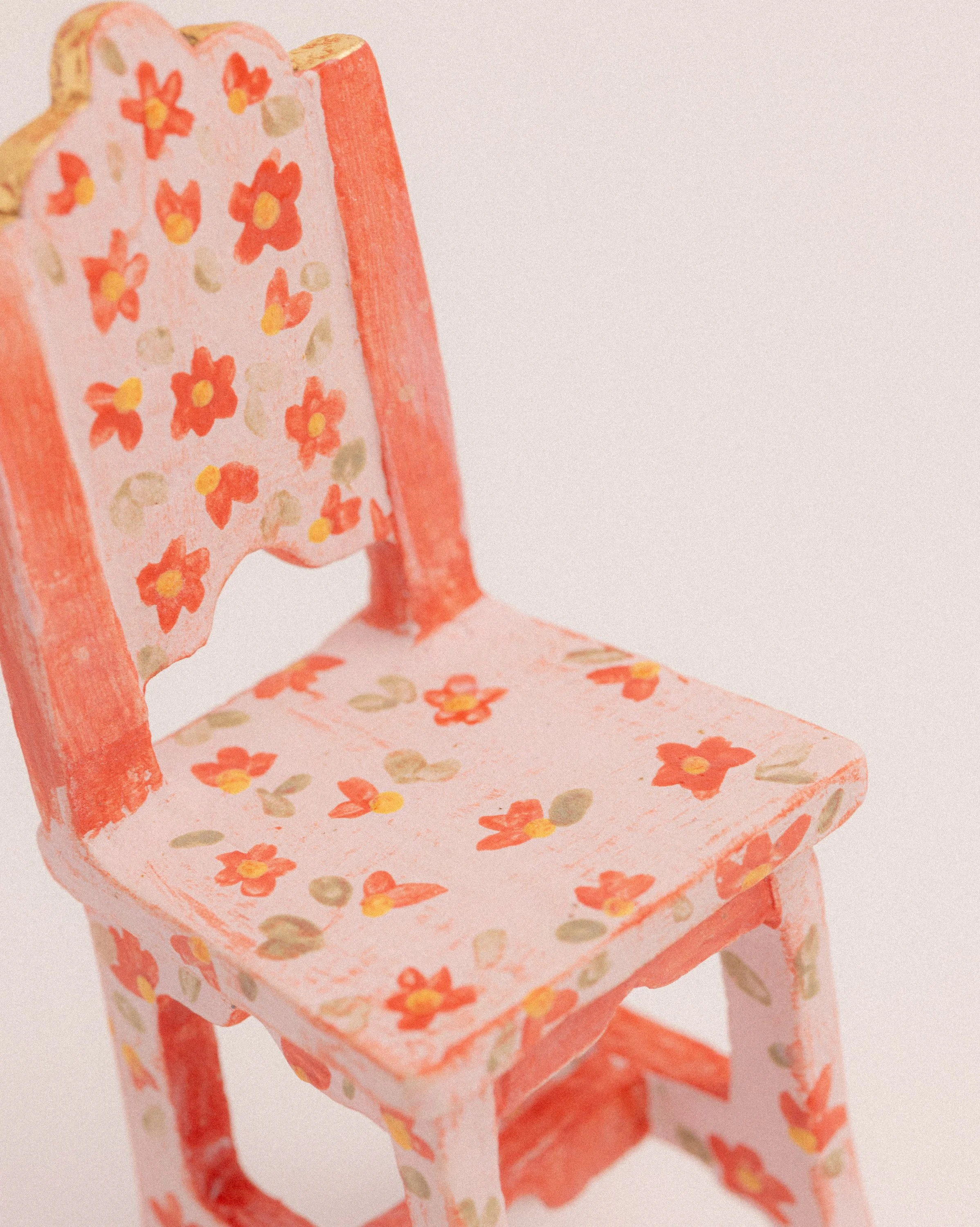 A small ceramic chair sculpture painted pink with a floral pattern featuring red and gold flowers and green leaves, shown from a side angle against a plain background.