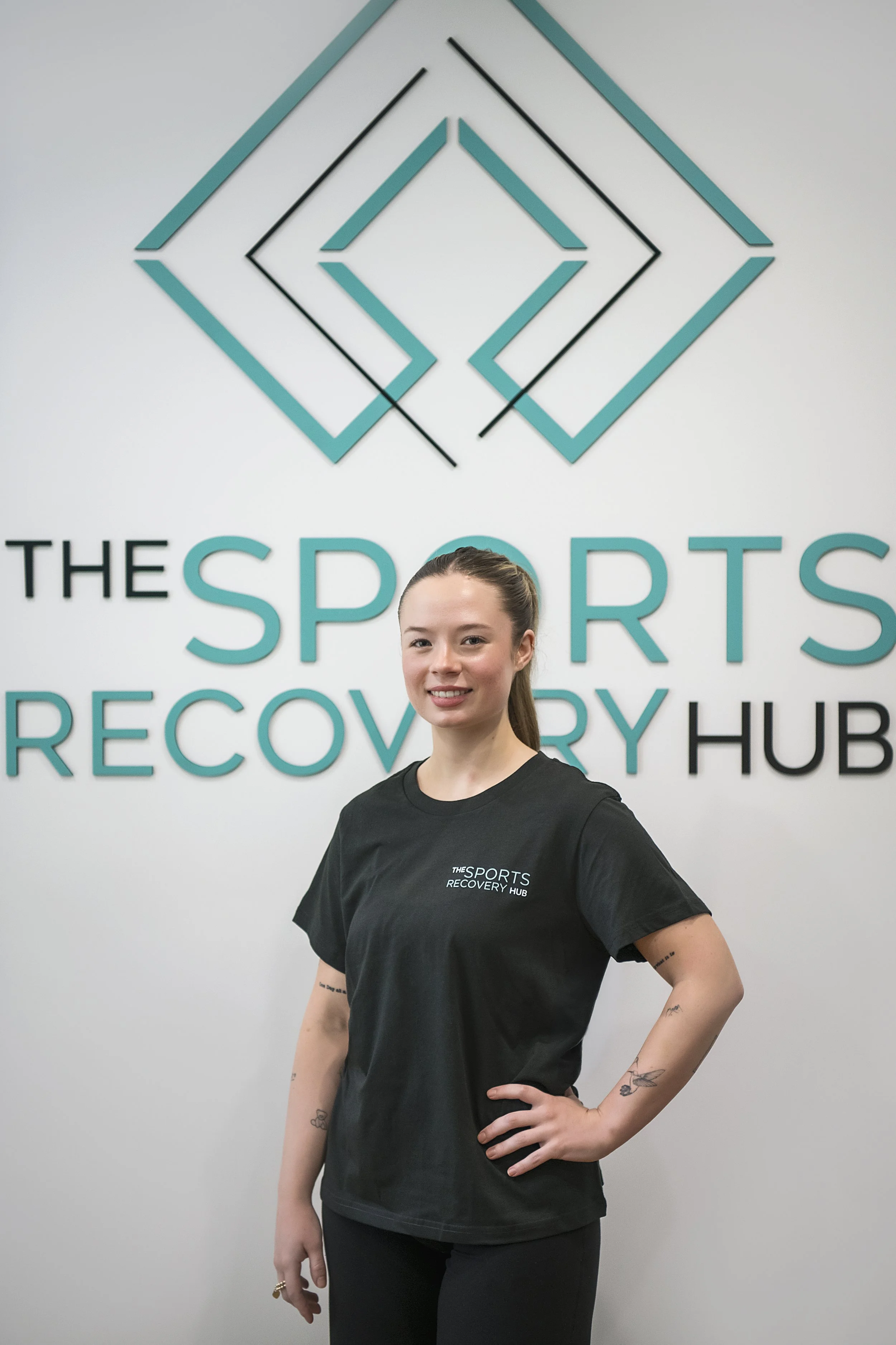 A young woman with long hair tied back, standing in front of a wall with the logo of the Sports Recovery Hub. She is wearing a black t-shirt with the same logo, smiling slightly, with her left hand on her hip and her right hand hanging down.