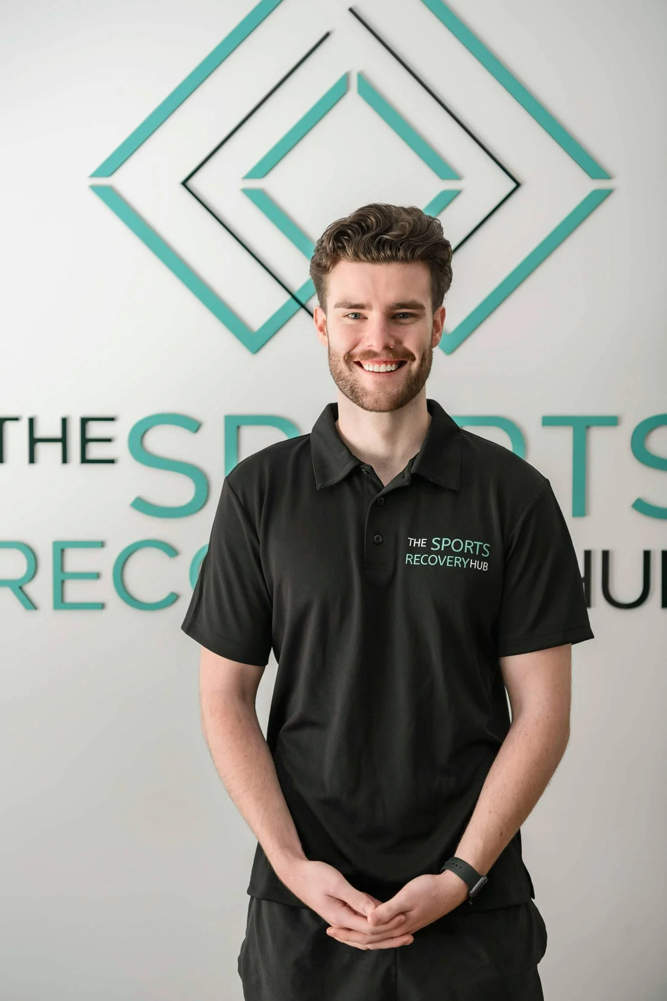 A young man with brown hair and a beard smiling while wearing a black polo shirt with the logo for The Sports Recovery Hub, standing in front of a wall with a geometric teal and black design and the text "THE SPORTS RECOVERY HUB".