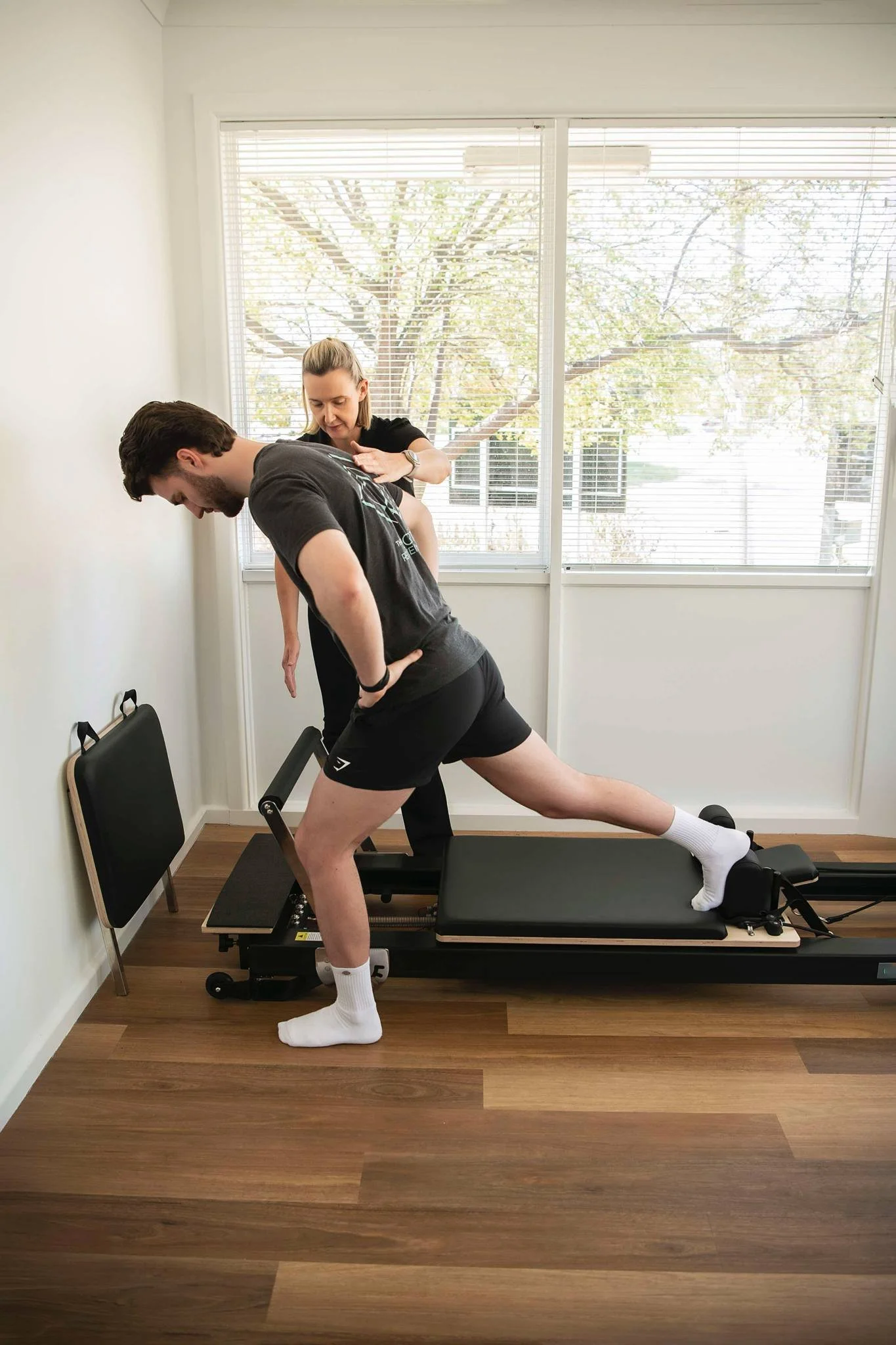 A man is exercising on a Pilates reformer machine while a woman presses on his back, assisting with his workout, clinical pilates for corrective rehab