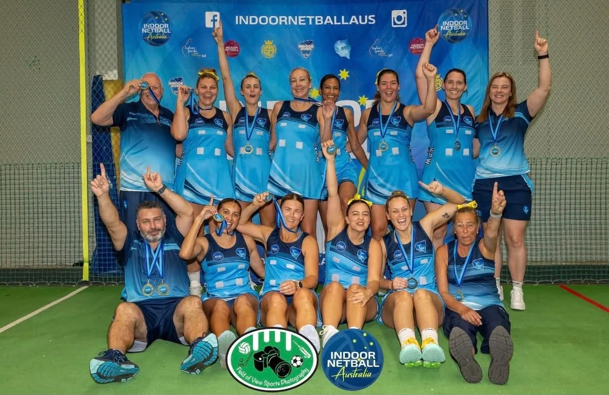 Group of female indoor netball players and coaches posing for a photo on a court, holding medals, thumbs up, and wearing matching blue sports uniforms, with a blue banner and sponsorship logos in the background.