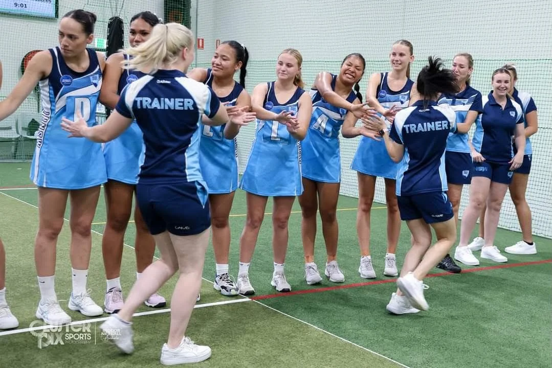 A group of female netball players in blue uniforms on an indoor court, celebrating and high-fiving each other during a game or practice, with a coach or trainer with back to camera in matching uniform.