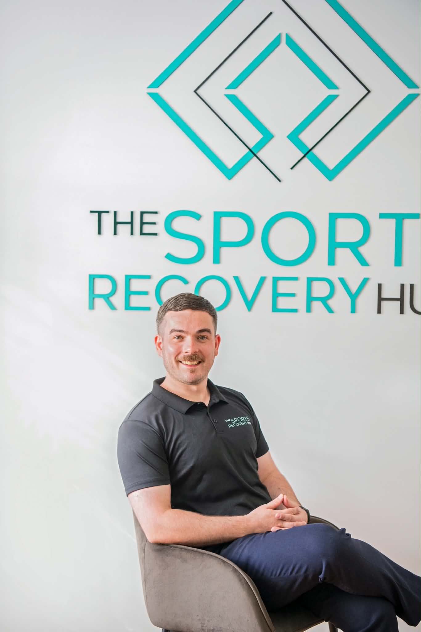 A man sitting on a chair in front of a wall with a logo that reads 'The Sport Recovery Hub' with geometric shapes in blue and black.