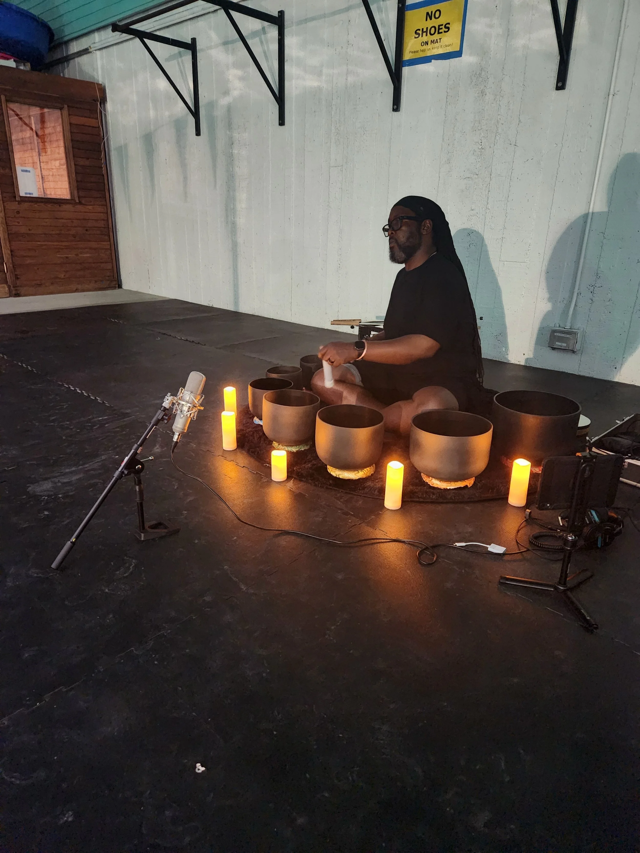 A person with long dreadlocks and glasses practicing sound healing with crystal bowls in a bright room.