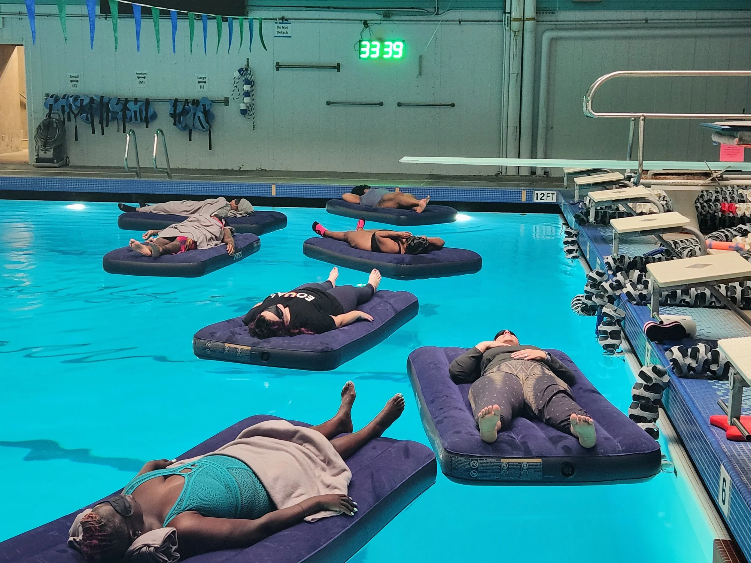 People relaxing on inflatable mattresses in a swimming pool at night, illuminated by candles, with a rocky wall, palm trees, and a building with large windows in the background.