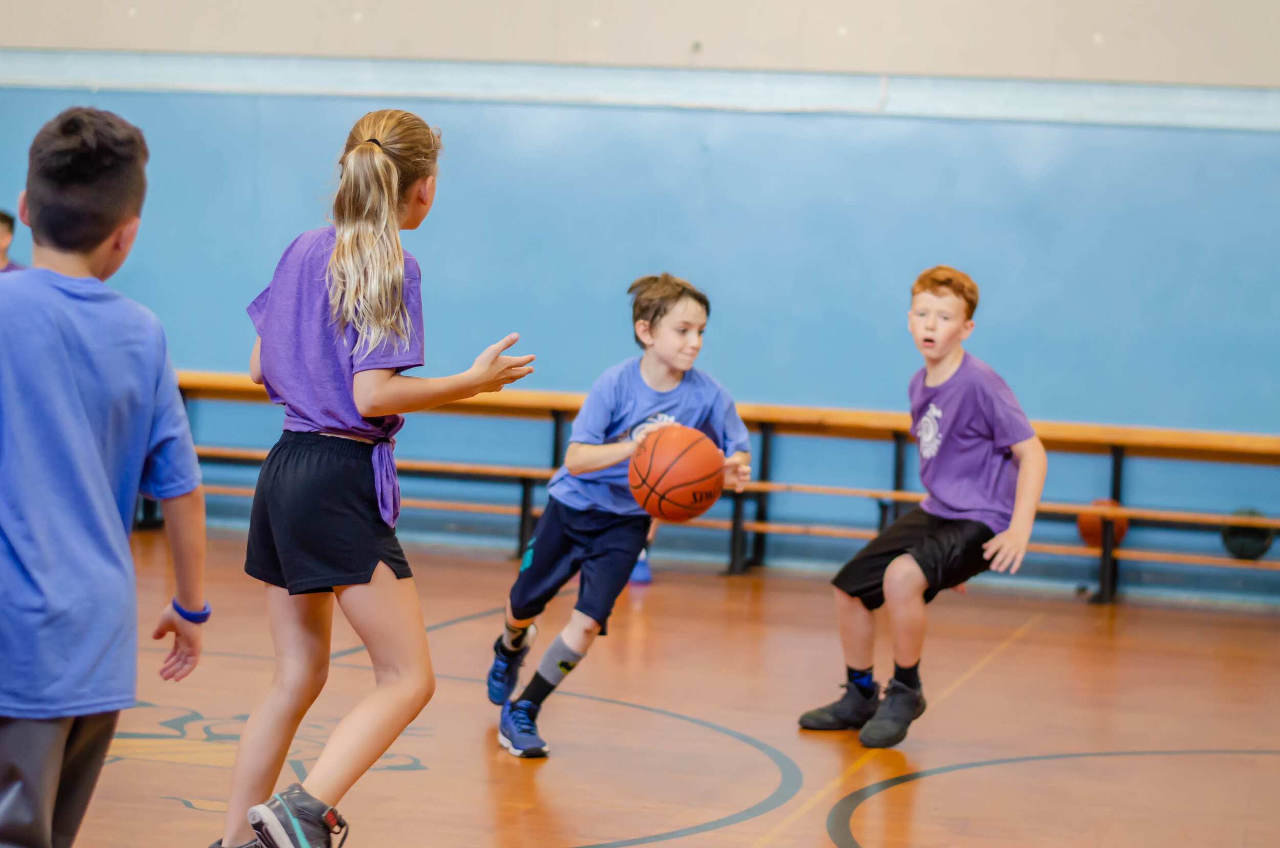 Children playing basketball in a gymnasium, some wearing purple shirts and one boy in a blue shirt dribbling a basketball.