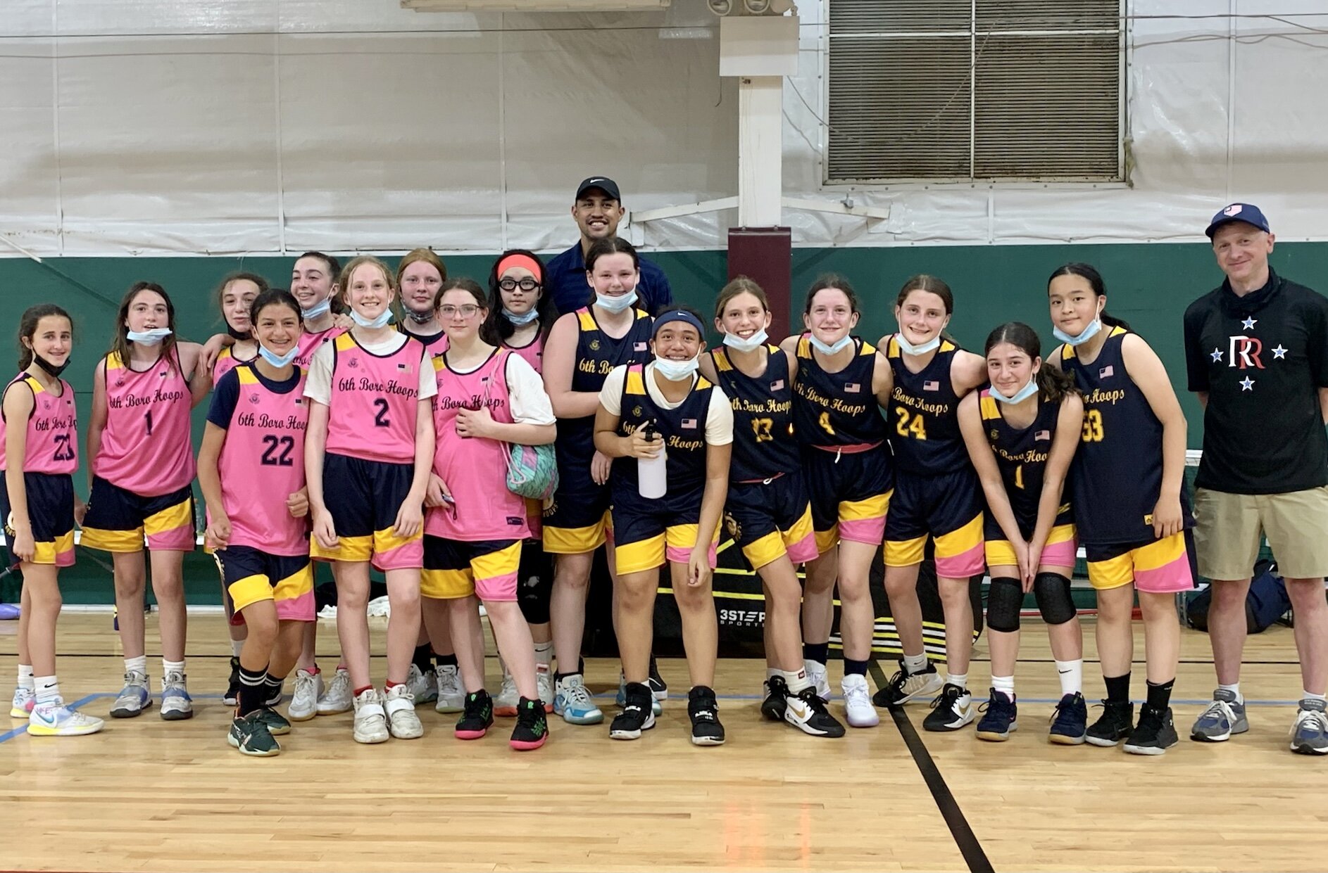A girls' basketball team standing in a gym, wearing matching pink and navy jerseys, with two coaches, all smiling.