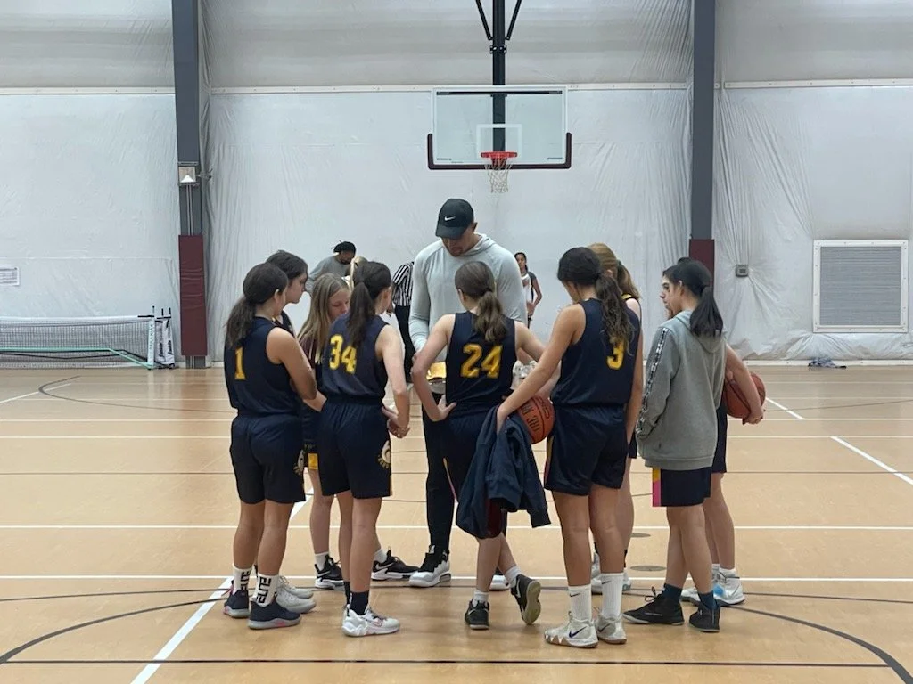 A girls basketball team in a huddle during a timeout on the court with their coach.