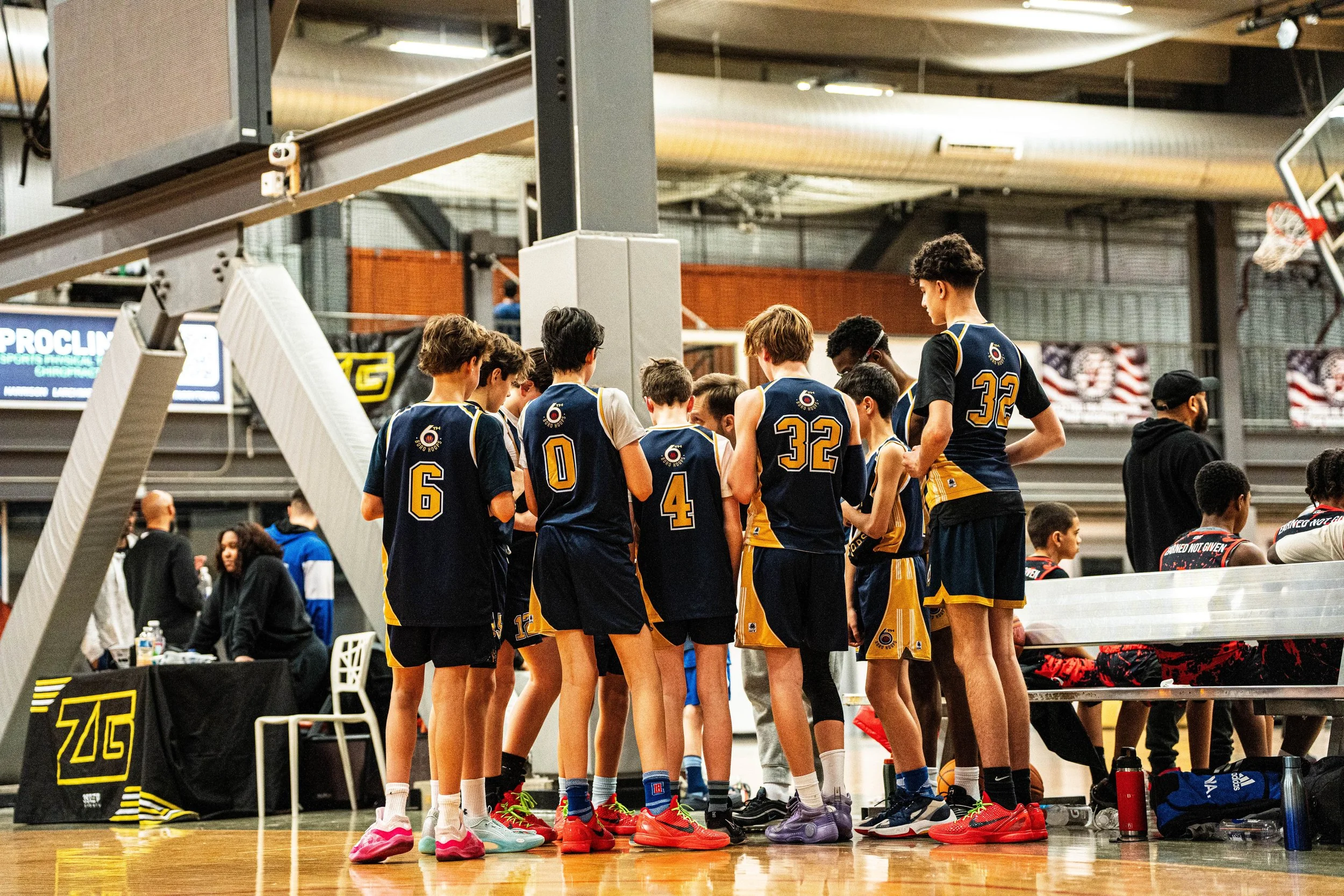 A group of young basketball players in navy and yellow jerseys huddle during a timeout on the court, with a basketball resting on the bench nearby.